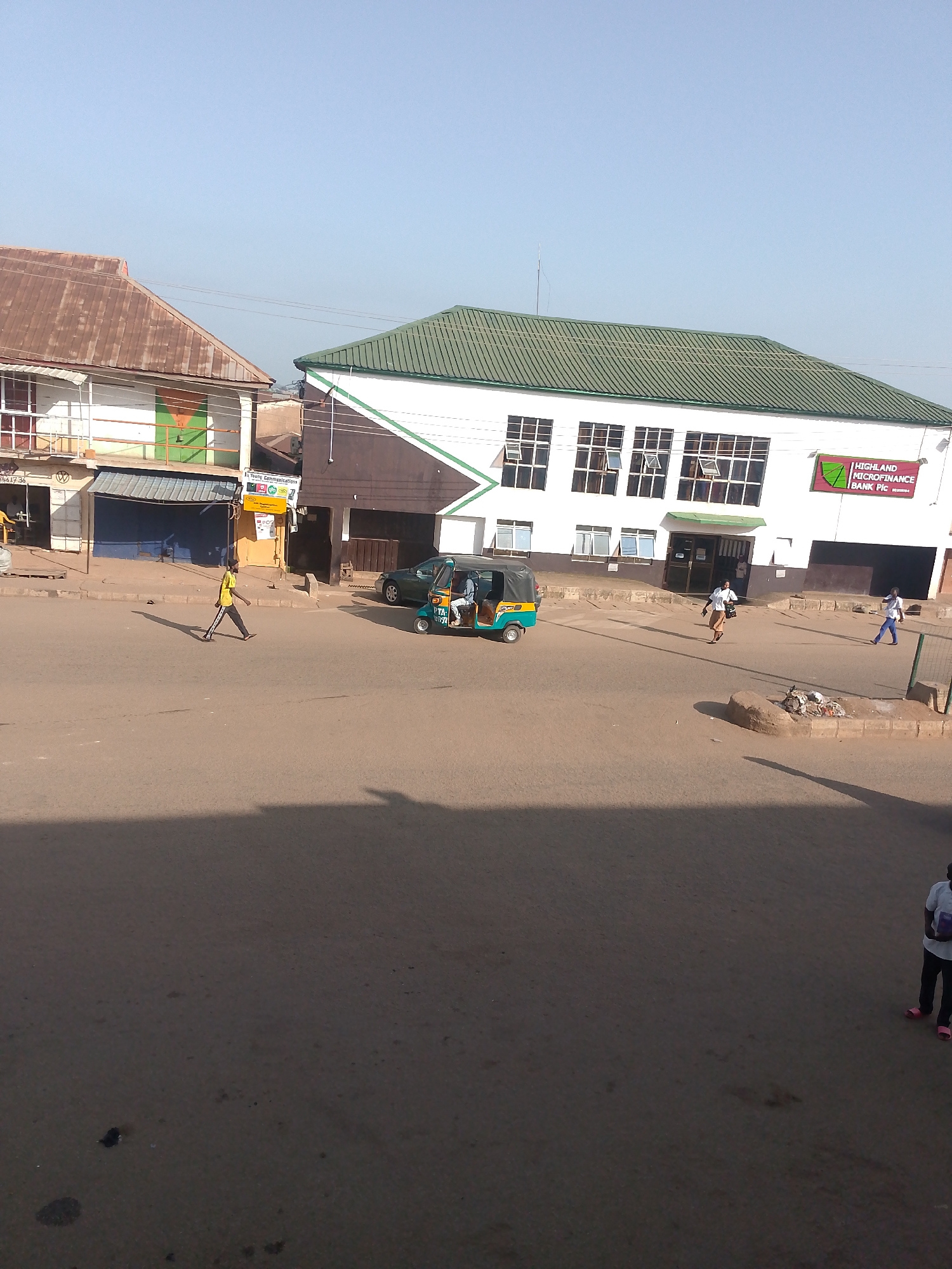 The image captures a street scene in Bukuru, Nigeria, on a clear, sunny day. In the foreground, a paved road is visible, with a significant shadow cast from an unseen object to the left, suggesting it is late afternoon or early morning.
Dominating the mid-ground are several buildings. On the left, a two-story building with a corrugated iron roof and white facade features shops with closed shutters and some open spaces. A person in a yellow shirt and dark pants walks across the road from left to right, casting a long shadow. To the right of this building is a modern, two-story structure with a green corrugated iron roof and a white facade, featuring numerous large windows. A prominent sign on this building reads "HIGHLAND MICROFINANCE BANK Plc". An entrance with glass doors is visible on the ground floor.
Several vehicles are present. A green tricycle with passengers is stopped in the road in front of the bank. A dark green car is parked on the right side of the road.
In the background, more buildings and a clear sky are visible. On the far right of the image, a person wearing a white shirt and dark pants stands facing away from the camera, holding a purple object. Another person in a white shirt and dark pants is walking to the right in front of the bank building.
The environment appears to be an urban commercial area, with the road serving as a thoroughfare for pedestrians and vehicles. The buildings suggest a mix of older and newer commercial establishments.