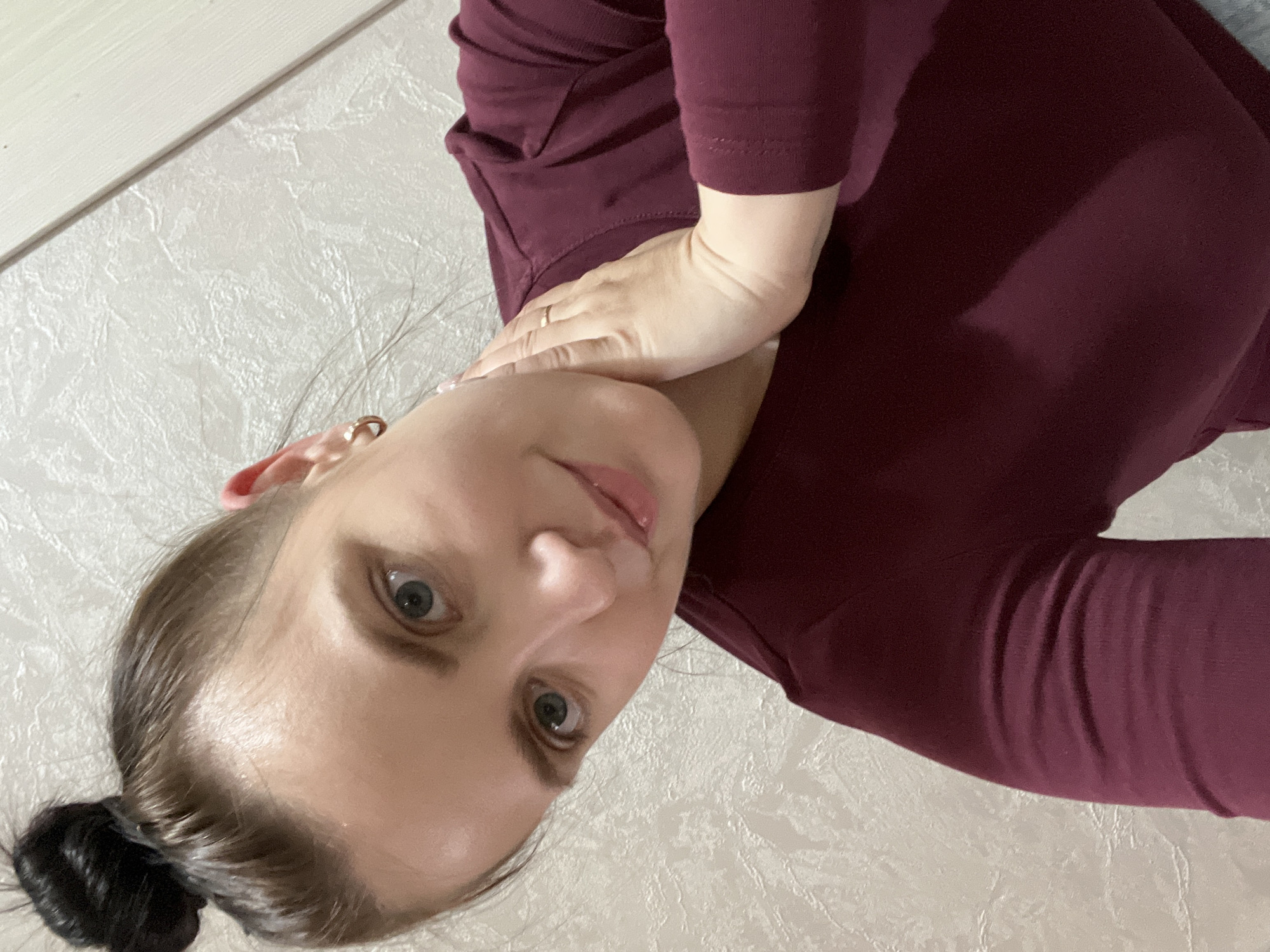 An indoor, vertically-oriented close-up portrait features a woman with her head tilted, looking directly at the camera. The setting appears to be a residential interior in Марковский, Russia.
The subject is a woman with light-colored eyes and dark hair pulled back into a bun. She wears small, golden hoop earrings. Her expression is neutral, with her eyes wide and a subtle hint of a smile on her lips. She is dressed in a dark maroon, long-sleeved top, which covers her torso and arms. Her right hand is visible, resting on her neck or upper chest area, and she wears a simple ring on her right ring finger.
The background consists of a light, possibly off-white, textured wall, likely wallpaper. A horizontal white strip, possibly a baseboard or door frame, is visible along the top edge of the image. The lighting is even, suggesting indoor artificial light or diffuse daylight. There is no visible text in the image. The overall impression is a candid, eye-level selfie.