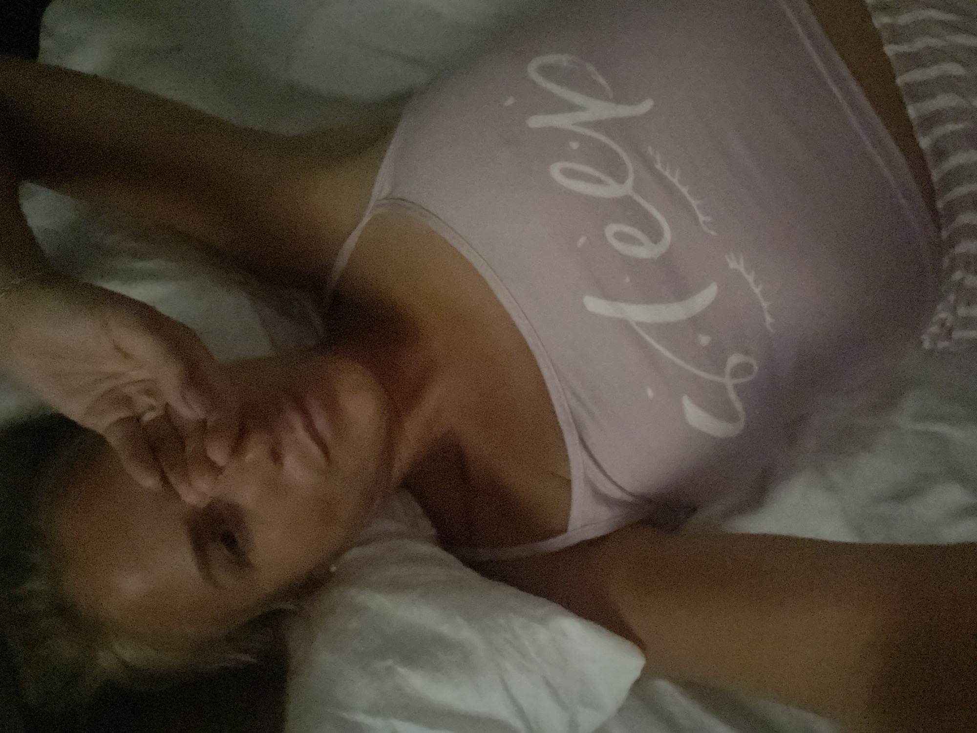 A woman is lying in bed, covering her eyes with her hand. She is wearing a light pink tank top with white lettering that says "sleep" and a drawing of eyelashes. Her hair is blonde and pulled back. The bed has white sheets and a white pillow. The image is taken from a high angle, looking down at the woman. The setting appears to be indoors, possibly a bedroom. There is no information about the location in Baranovka, Russia.