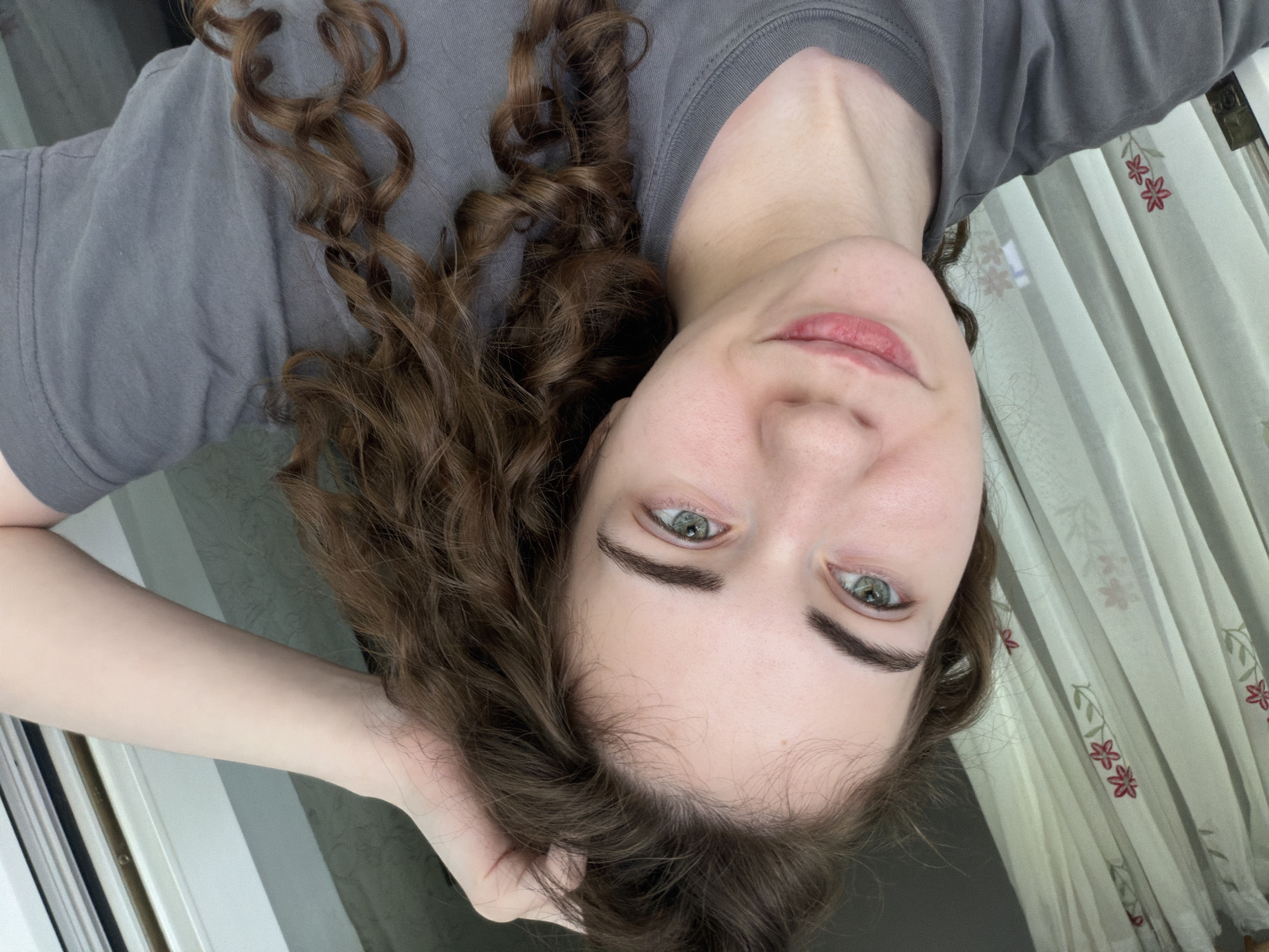 An over-the-shoulder, medium shot shows a young woman with curly brown hair and fair skin, looking slightly to her left. She is wearing a grey t-shirt and has her right hand raised, touching her hair. Her eyes are a striking light green, and her lips are slightly parted. Behind her, sheer white curtains with a floral pattern are visible. The lighting suggests an indoor setting, possibly near a window. There are no identifiable landmarks or context to place this image specifically within Zavodoukovsk, Russia.