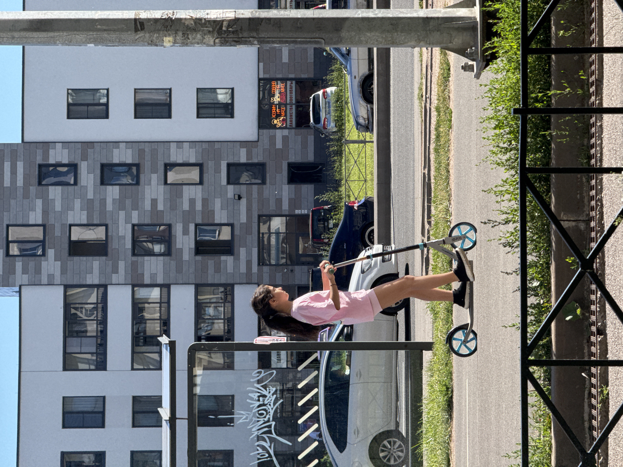 A young girl, likely a teenager, is captured in a vertical, low-angle shot while riding a scooter. She is dressed in a pink t-shirt and pink shorts, with her dark hair tied back in a ponytail. The girl is looking upwards and to her right, with a slight smile suggesting a moment of enjoyment or anticipation. She is wearing dark socks and black sneakers.
The scooter is grey with blue wheels. The girl's legs are extended, and she appears to be in motion, pushing off with her right foot. Her left foot is on the scooter's deck.
The scene is set on a paved sidewalk next to a road. In the background, a modern multi-story building with a grey and white facade dominates the view. The building features numerous windows, some reflecting the surroundings. A few cars are visible on the road, including a silver hatchback and a darker vehicle. To the right of the road, a patch of green grass and foliage lines the sidewalk, bordered by a dark metal fence. Sunlight casts shadows, indicating it is daytime. The overall atmosphere seems to be a bright, sunny day in an urban environment.
Visible text includes what appears to be Russian lettering on a sign on the building facade, which reads "ХОЗЯИН СОБАКА" and "ЛАКОМСТВА ЗООТОВАРЫ", suggesting a pet store or related business. There is also graffiti on a glass structure in the foreground. The overall impression is a candid moment of a girl enjoying an outdoor activity in a residential or commercial area of Saint Petersburg, Russia.