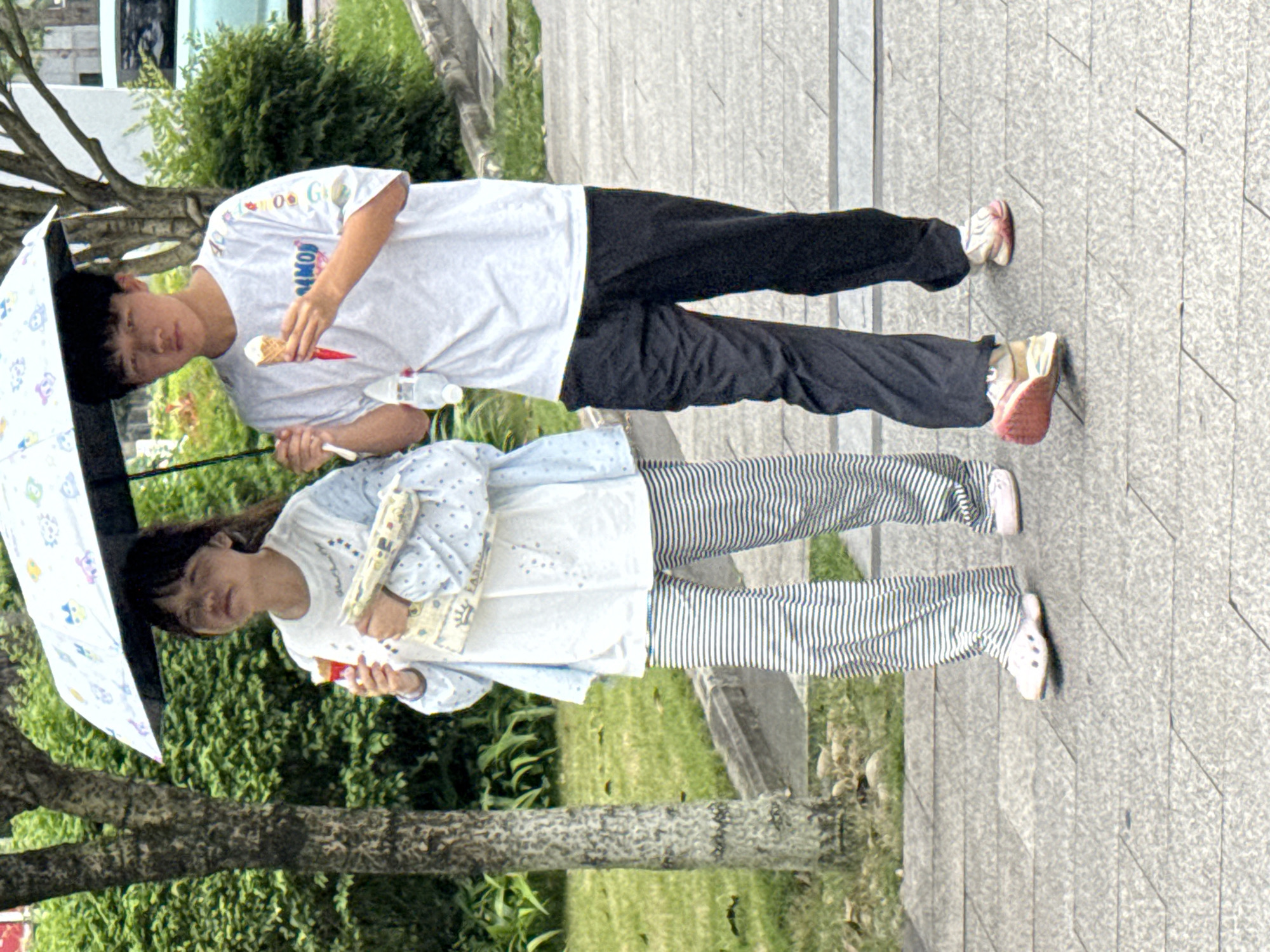 Two young individuals stand together on a paved walkway, with greenery and a tree visible in the background. The individual on the left holds an umbrella and wears a patterned shirt with striped pants. They are holding a colorful item, possibly a wrapper or snack. The individual on the right wears a white t-shirt, black pants, and holds an ice cream cone. A water bottle is also visible next to them. The location is Jiangjin, China.