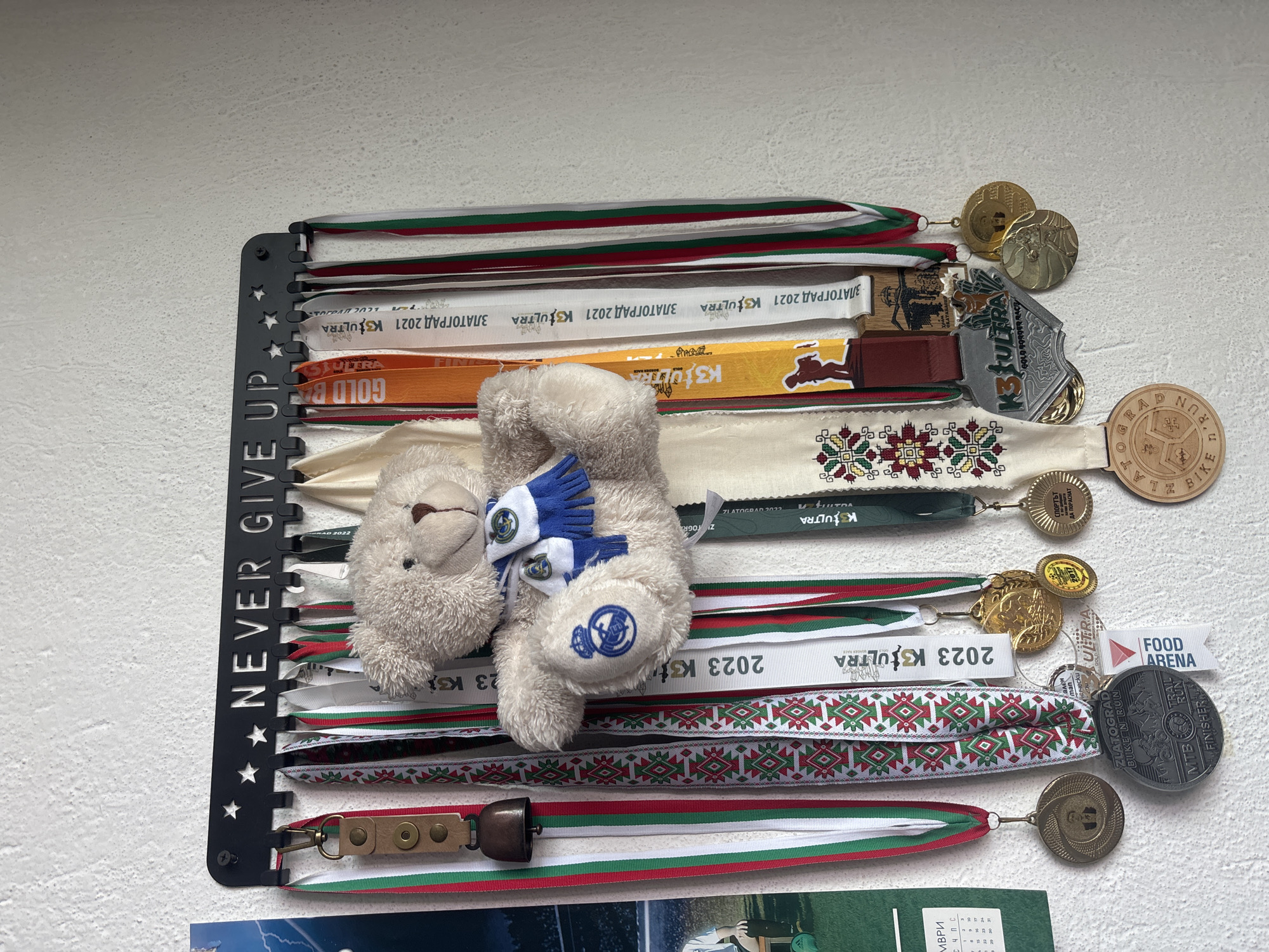 The image displays a collection of athletic medals and their ribbons, neatly hung on a black metal medal hanger against a white, textured wall. The medal hanger is rectangular and features cut-out stars on its left side, with the phrase "NEVER GIVE UP" running vertically along its edge.
Numerous ribbons, predominantly in white, green, and red, are draped over the hooks of the hanger. Many ribbons feature traditional Bulgarian patterns in these colors. Among the ribbons are various medals, including circular metallic ones in gold, silver, and bronze tones, and one distinctive wooden medal. A small, light brown plush teddy bear, wearing a blue and white striped scarf and matching gloves, sits atop some of the ribbons in the center of the display. The teddy bear's paw has an emblem stitched onto it.
Several pieces of text are visible on the ribbons and medals, mostly related to sports events. Repeated inscriptions include "ЗЛАТОГРАД 2021 K3ULTRA" and "2023 K3ULTRA", referencing races in Zlatograd. Other identifiable texts include "ZLATOGRAD RUN 'n' BIKE" on the wooden medal, "MTB TRAIL RUN FINISHER", "GOLD AGROER RACE", and "FOOD ARENA" on a small tag. Another ribbon displays "СПОРТЪТ ДА ПОРАСНАТ", which translates to "SPORT TO GROW". Part of an acronym, "МВРИ", is visible at the bottom left. The display suggests a passion for endurance sports and commemorates past achievements, likely from events held in the Zlatograd region of Bulgaria.