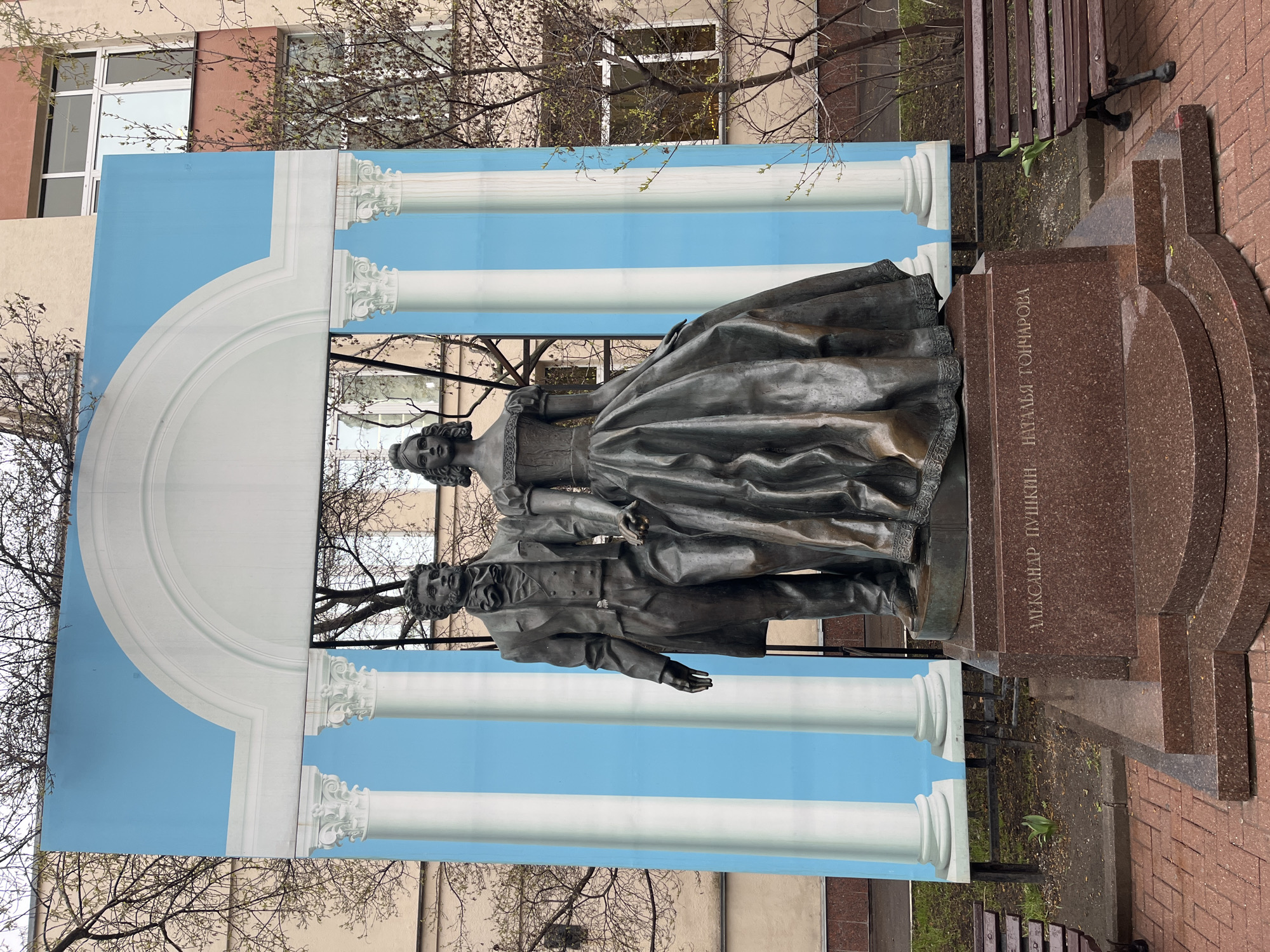 The image captures a statue of Alexander Pushkin and Natalia Goncharova, located in Moscow, Russia. The bronze statue depicts the couple standing side-by-side. A backdrop featuring a classical architectural design of white columns against a blue sky is placed behind the statue. The statue stands on a reddish-brown stone base with engraved inscriptions. The surrounding environment includes a park setting with bare trees, a building facade in the background, a park bench on the right, and brick pavement around the statue.