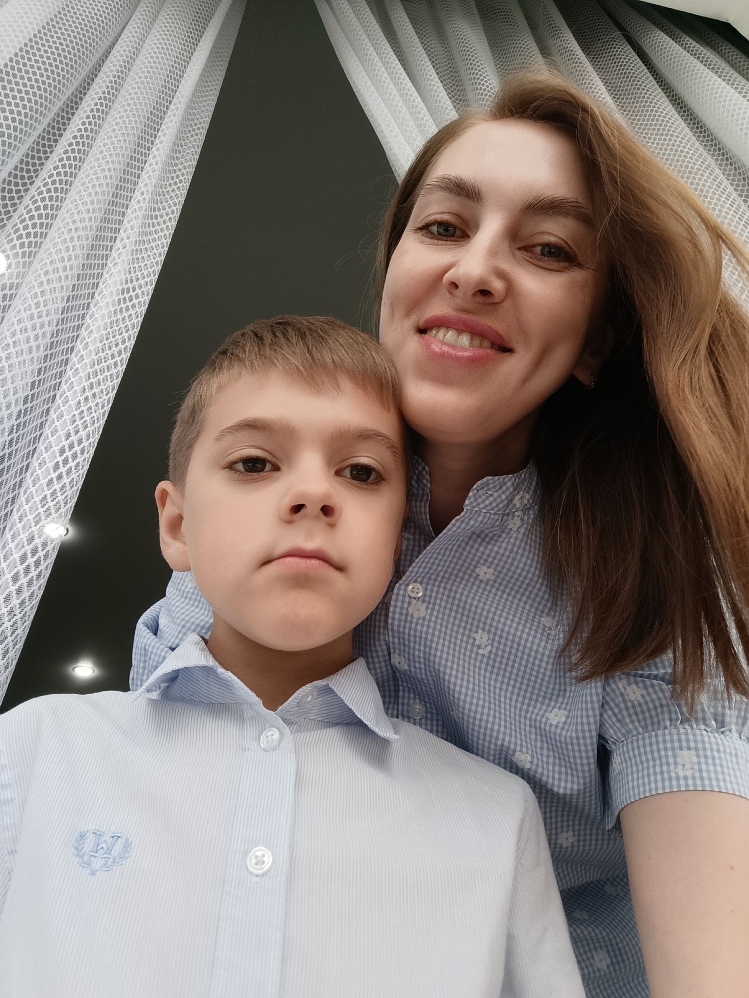 A woman and a boy are posing indoors. The woman is fair-skinned with brown hair, smiling at the camera, and wearing a blue and white patterned collared shirt. The boy is fair-skinned with short brown hair and a serious expression. He's wearing a light blue collared shirt with a logo on the left side. A sheer white curtain is visible in the background, with dark walls behind it. The location is Chaikovsky, Russia.