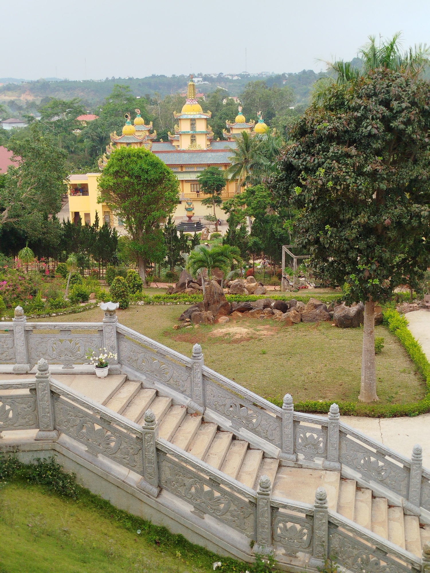 The image depicts a multi-tiered ornate stone staircase in Phú An, Vietnam. The staircase leads down into a well-maintained park-like setting, featuring trees, rocks, and manicured grass. The background reveals a multi-story yellow building with distinct domes and tiered roofs, typical of Buddhist temple architecture. The sky is overcast, creating soft light across the scene.