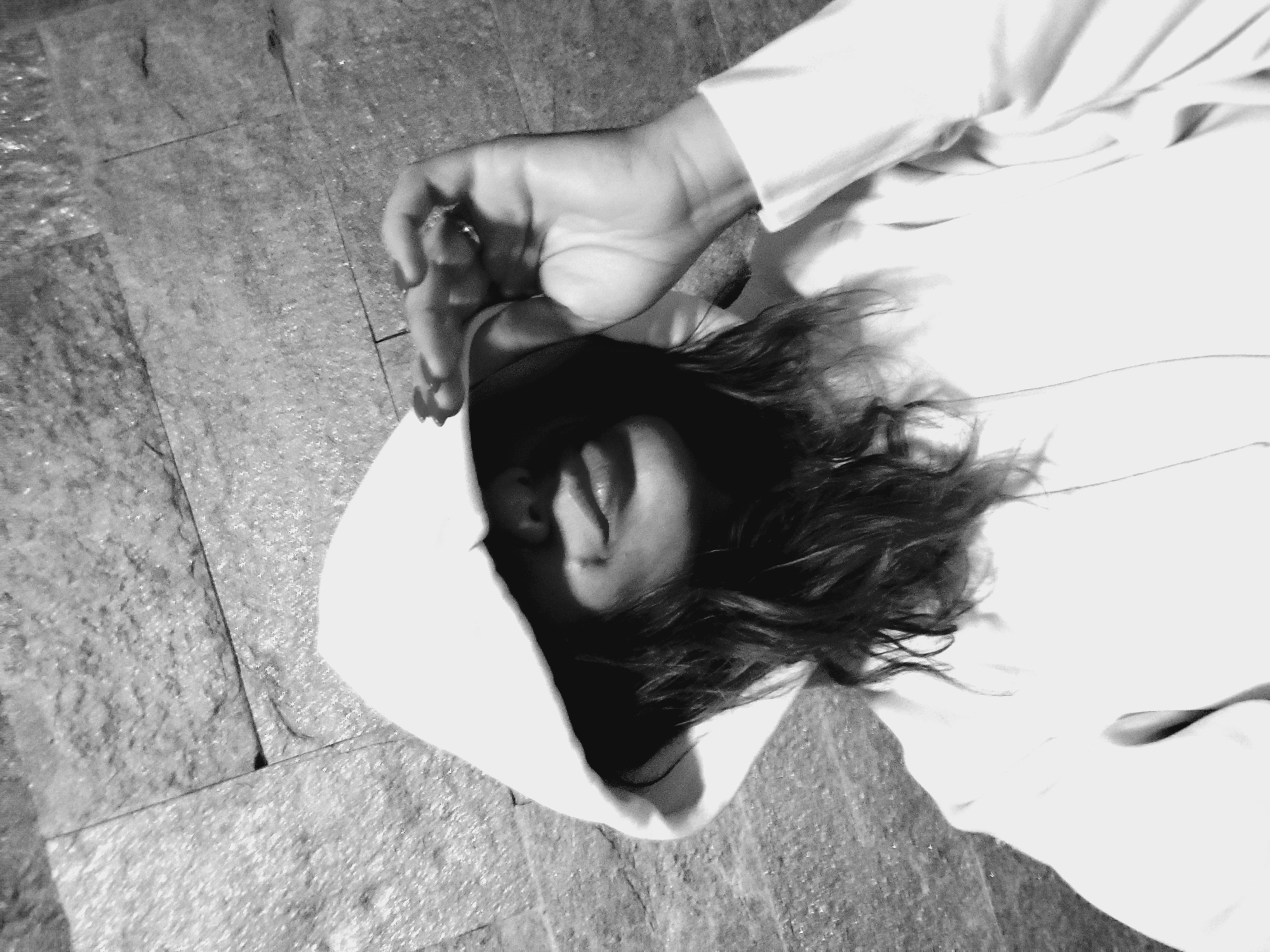 A black and white, medium close-up, low-angle selfie shows a person wearing a white hooded sweatshirt, with the hood pulled up to obscure most of their face. The person's mouth is slightly parted, with their lips slightly pursed, and their dark, wavy hair is visible beneath the hood. Their right hand is raised to the hood, with fingers curled and manicured nails visible. The background consists of a textured, stone-tiled wall. The image was taken in the city of Sozopol, Bulgaria.