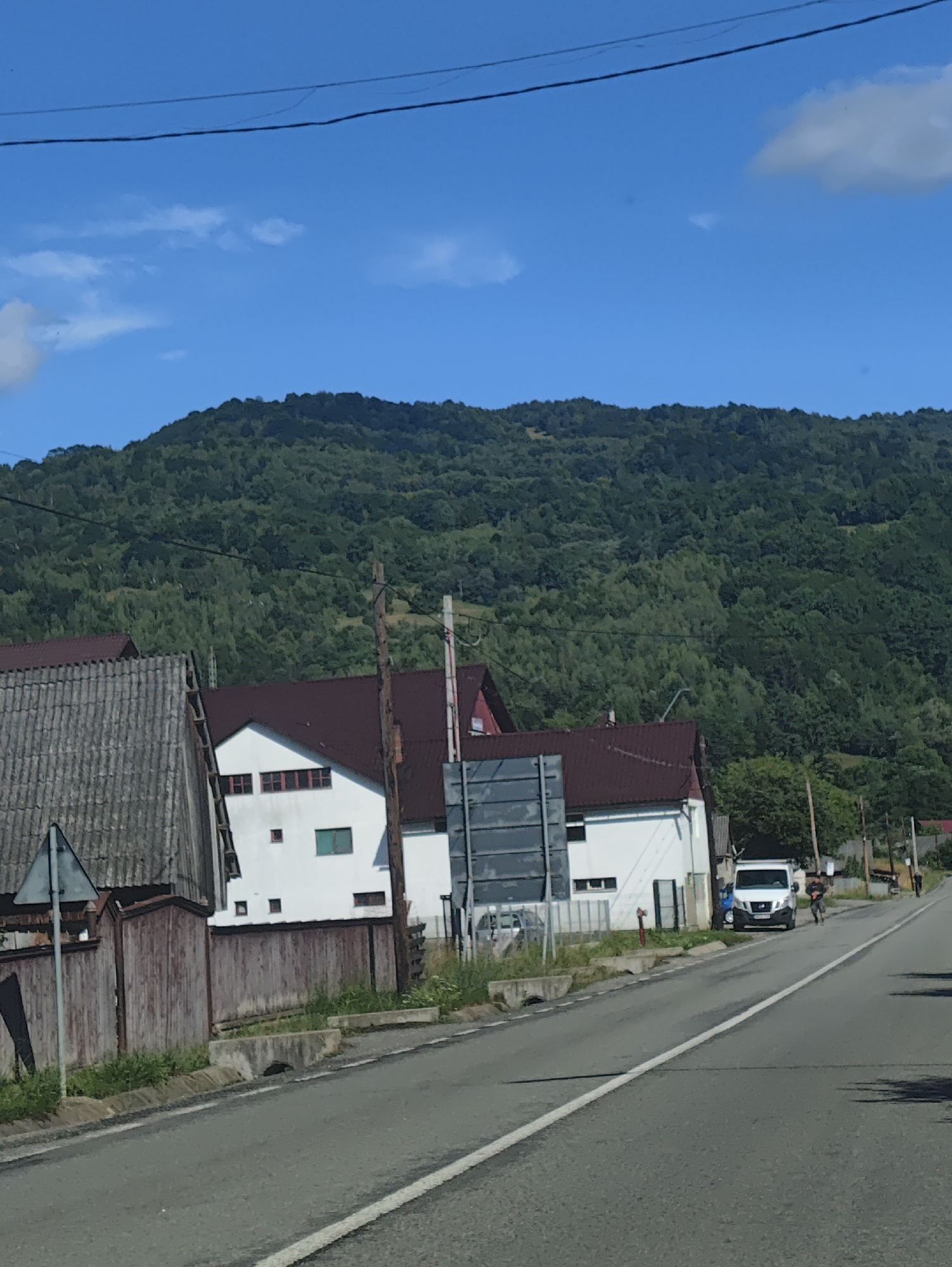 The image depicts a rural scene in Leordina, Romania, with a paved road running through a village. On the left side of the road, there is a rustic wooden fence and a weathered, grey-roofed building. Adjacent to this is a white building with a dark red roof, featuring several windows. A white van is parked on the right side of the road, with two individuals standing near it. Further down the road, more houses and trees are visible, all set against a backdrop of a lush, green, tree-covered mountainside. The sky above is a clear blue with a few scattered clouds. The overall impression is that of a quiet, rural community on a sunny day.