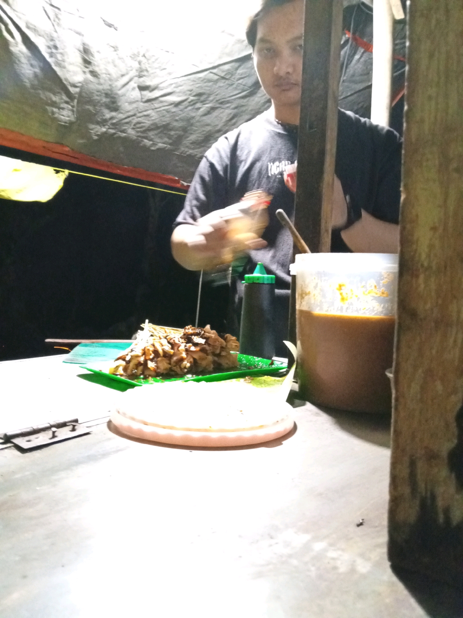 In Kedungwinong, Indonesia, a person stands behind a makeshift food stall, preparing a dish of satay. The stall appears to be set up outdoors, possibly under a tarp or canopy, with natural light and artificial lighting contributing to the scene's illumination. The person, wearing a dark t-shirt, is in the process of serving food, with their hands blurred due to motion. In front of them, a plate holds a portion of satay, accompanied by what appears to be a dipping sauce in a clear plastic container. Other items visible include a squeeze bottle with a green cap and a white plate or serving dish. The overall setting suggests a casual dining environment, common in street food establishments.