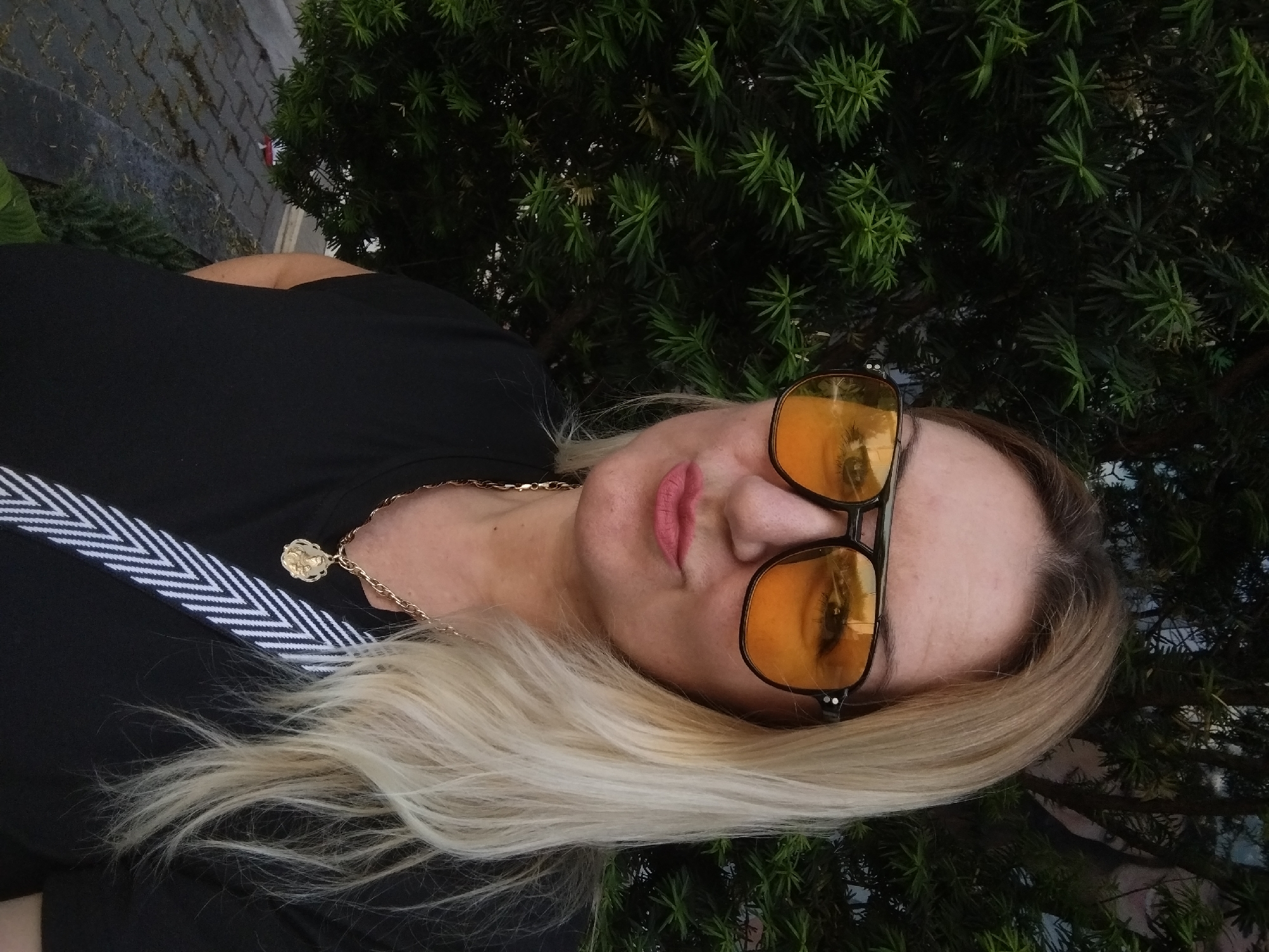 A woman with blonde hair is wearing yellow sunglasses, a black shirt with a white and black striped detail, and a gold necklace. She is outdoors, with evergreen foliage visible behind her. Based on the presence of distinctive evergreen species and the architectural style, it's likely the photo was taken in a park or garden within the city of Stara Zagora, Bulgaria. The woman is looking at the camera.