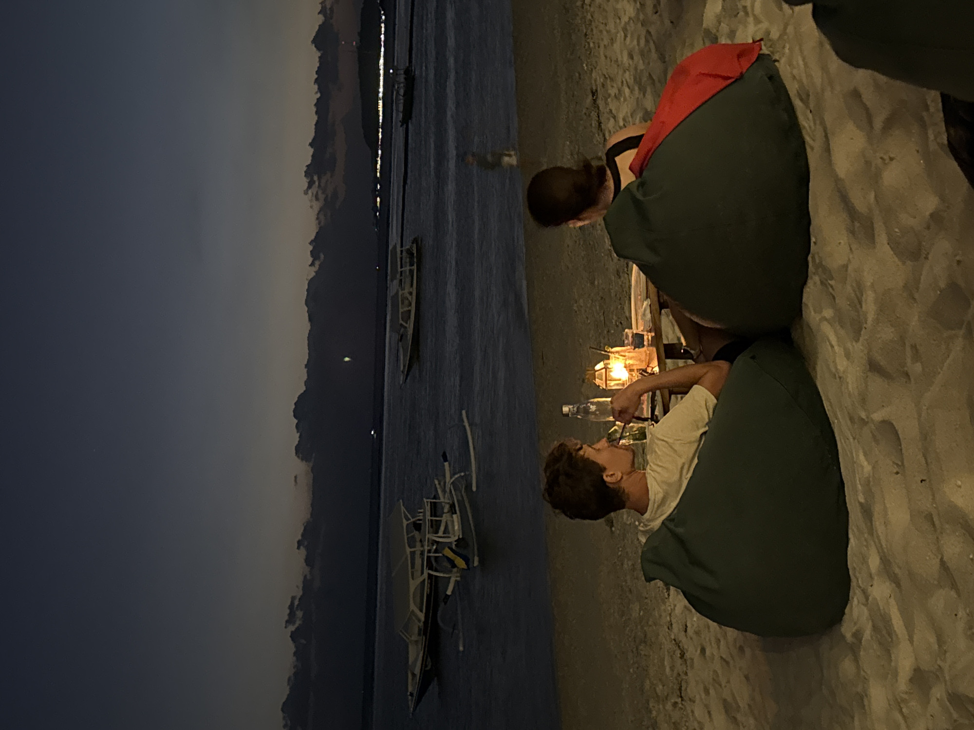 Two people, a man and a woman, are seated on beanbag chairs on a sandy beach at dusk. They are facing away from the camera, looking out at the ocean. A low table between them holds a lantern, a bottle of water, and two glasses. Several boats are anchored in the calm water offshore. The sky is dark blue with hints of color from the setting sun, and the clouds are dark and dramatic. The overall atmosphere is peaceful and romantic. The location is likely a beachside restaurant or bar in Indonesia, possibly on an island such as Bali.