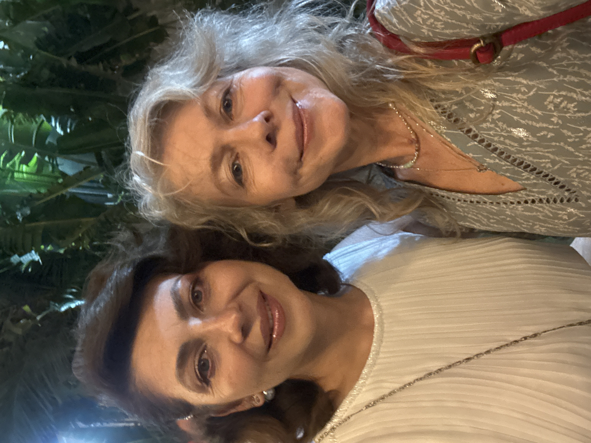 The image shows a close-up selfie of two women. The woman on the left has dark, wavy hair and is wearing a white, pleated top with a silver chain necklace. She has on earrings and appears to be wearing makeup. The woman on the right has lighter, wavy hair and is wearing a light blue patterned dress with a red strap over her shoulder. She is also wearing a necklace. Both women are smiling at the camera. The background appears to be outdoors with lush green foliage, suggesting a warm climate. The lighting is dim, consistent with evening or nighttime. The image was taken in Migdal HaEmek, Israel.