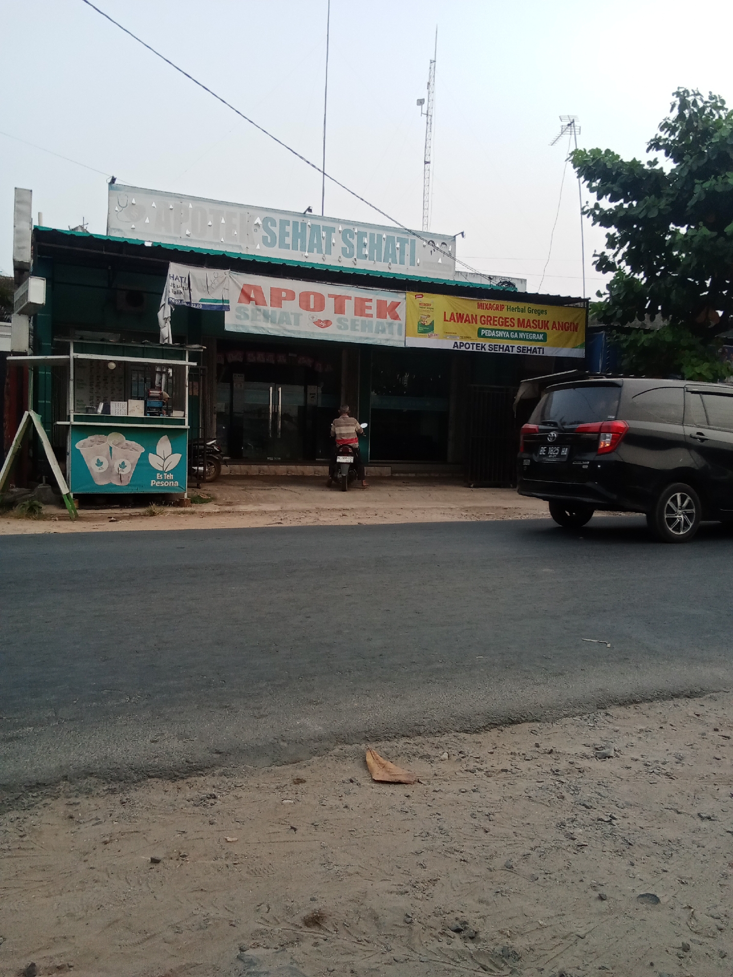 In the city of Toto Katon, Indonesia, a black Toyota Avanza, license plate BE 3825 HA, is parked in front of a pharmacy. The pharmacy has a prominent sign reading "APOTEK SEHAT SEHATI" in large red and blue letters. A smaller banner above the entrance advertises "MIXAGRIP Herbal Greget Lawan Greget Masuk Angin," indicating it sells medicine for cold and flu symptoms. To the left of the pharmacy is a small kiosk selling "Es Teh Pesona," or "Charming Iced Tea," with graphics of iced tea cups. A person on a motorcycle, wearing a striped shirt, is seen in front of the pharmacy. The street in front of the establishment is paved, with a sandy shoulder to the right. In the background, a tall radio tower and trees are visible against a clear sky.