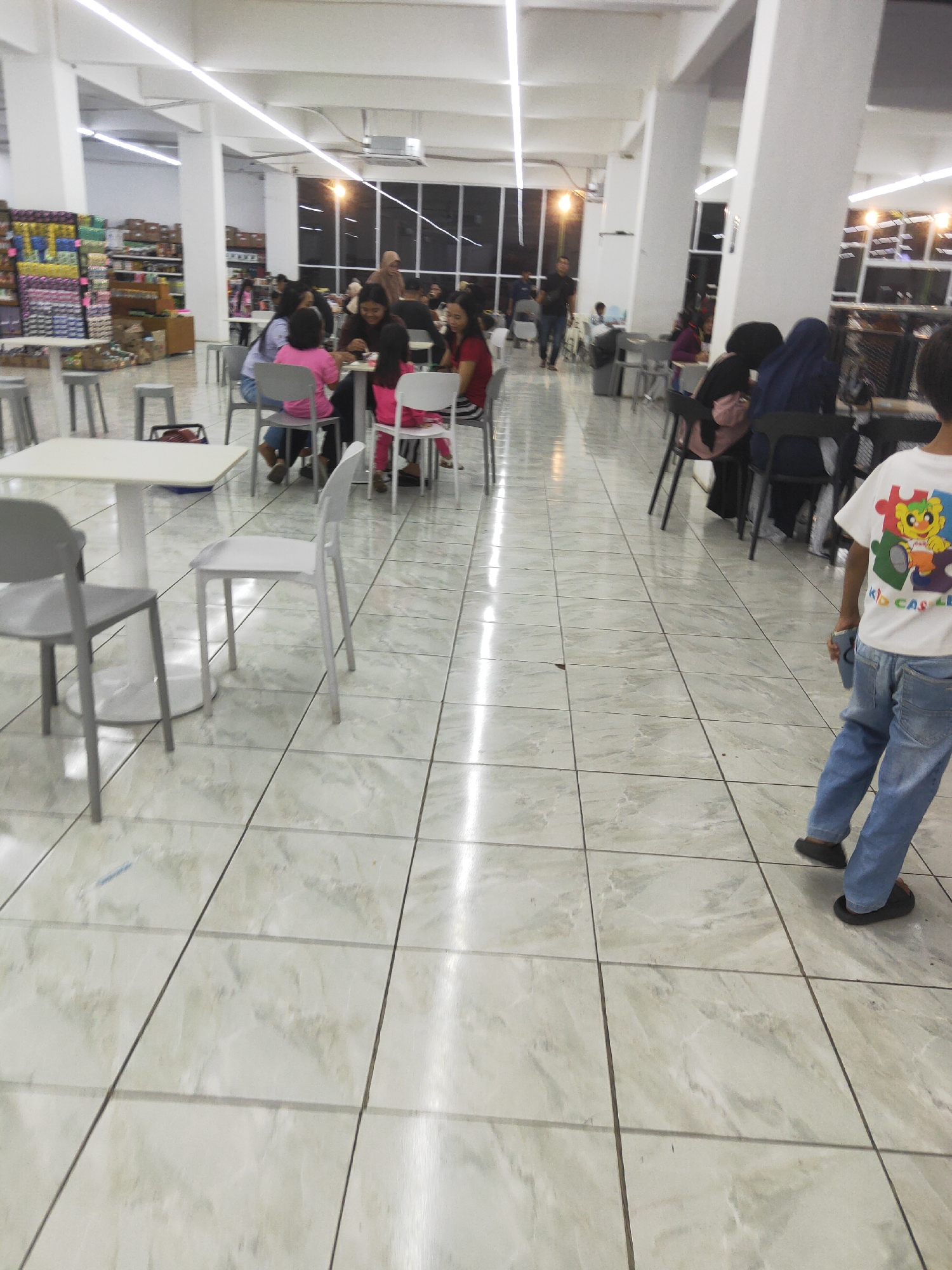The image depicts the interior of a food court in Sintang, Indonesia. The space is brightly lit by fluorescent ceiling lights and appears to be part of a larger establishment, possibly a supermarket or mall, given the shelving units visible in the background on the left. The floor is tiled with large, light-colored marble-patterned tiles.
Several tables and chairs are arranged throughout the food court. In the foreground, to the left, a white table with a pedestal base is surrounded by gray chairs. Further back, groups of people are seated at tables, eating and socializing. On the left side, a group of children and adults are gathered around a table. One child is wearing a pink shirt, and another is wearing a bright pink top. Further into the food court, several individuals are seated at tables with black chairs. Some of these individuals are wearing headscarves, indicating a cultural context.
In the right foreground, a young boy is walking away from the camera. He is wearing blue jeans and a white t-shirt with a colorful cartoon graphic and text that reads "KID CASTLE". He is wearing dark sandals.
The overall environment suggests a casual and busy dining area. The presence of multiple tables and chairs, along with people engaged in eating, points to a functioning food court. The store shelves in the background indicate that this food court is likely situated within or adjacent to a retail space.