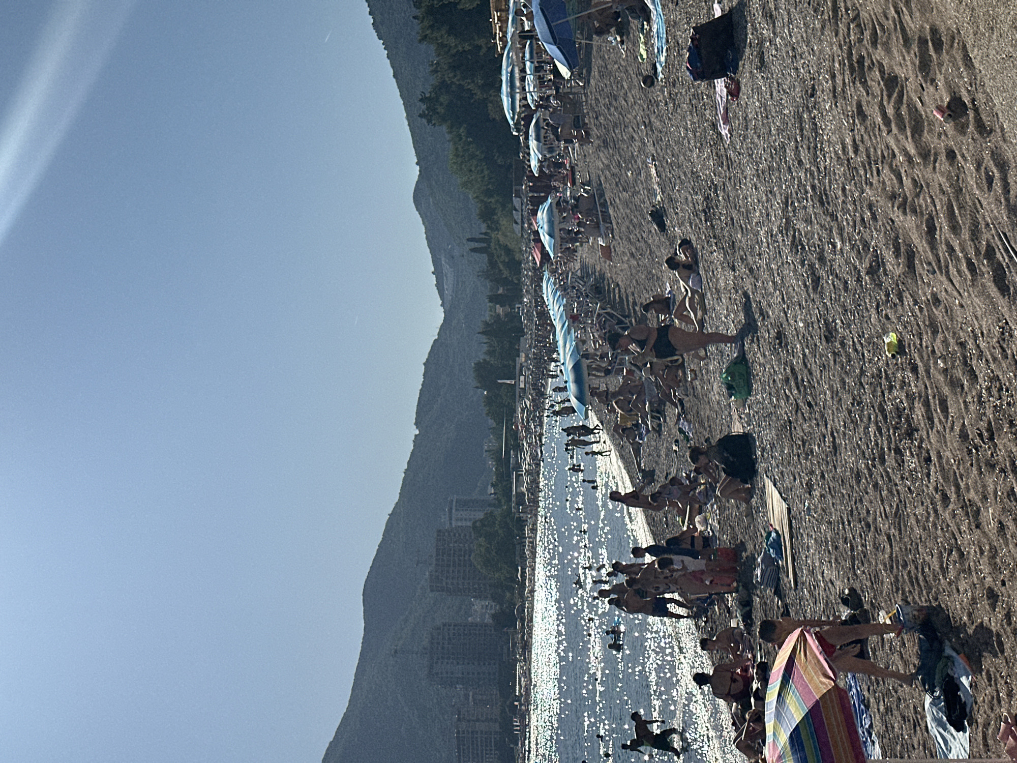 This image depicts a highly crowded beach scene in Budva, Montenegro, under clear, bright blue skies with intense sunlight. A range of tree-covered mountains forms the backdrop, with several multi-story buildings visible at their base, particularly on the left side of the bay. The body of water, likely the Adriatic Sea, occupies the left midground and foreground, intensely shimmering with numerous bright sun reflections, making details of individuals swimming or wading within it largely silhouetted.
The beach itself is composed of dark sand or fine pebbles and is densely populated with people. Numerous beach umbrellas, predominantly blue and white striped, are set up in rows, providing shade, with a distinctive multi-colored striped umbrella visible in the immediate foreground on the left. Many individuals are lying on sun loungers or towels on the sand, sunbathing or relaxing. Others are standing, walking, or engaging in various beach activities. In the foreground, near the multi-colored umbrella, a child stands beside a sand bucket and spade. Two empty beverage cans, one yellow and one red, are visible on the sandy surface in the far foreground, indicating litter. A dark bag and a beach ball are also present on the sand. The strong sunlight creates distinct shadows and a visible lens flare effect in the upper right quadrant of the sky.