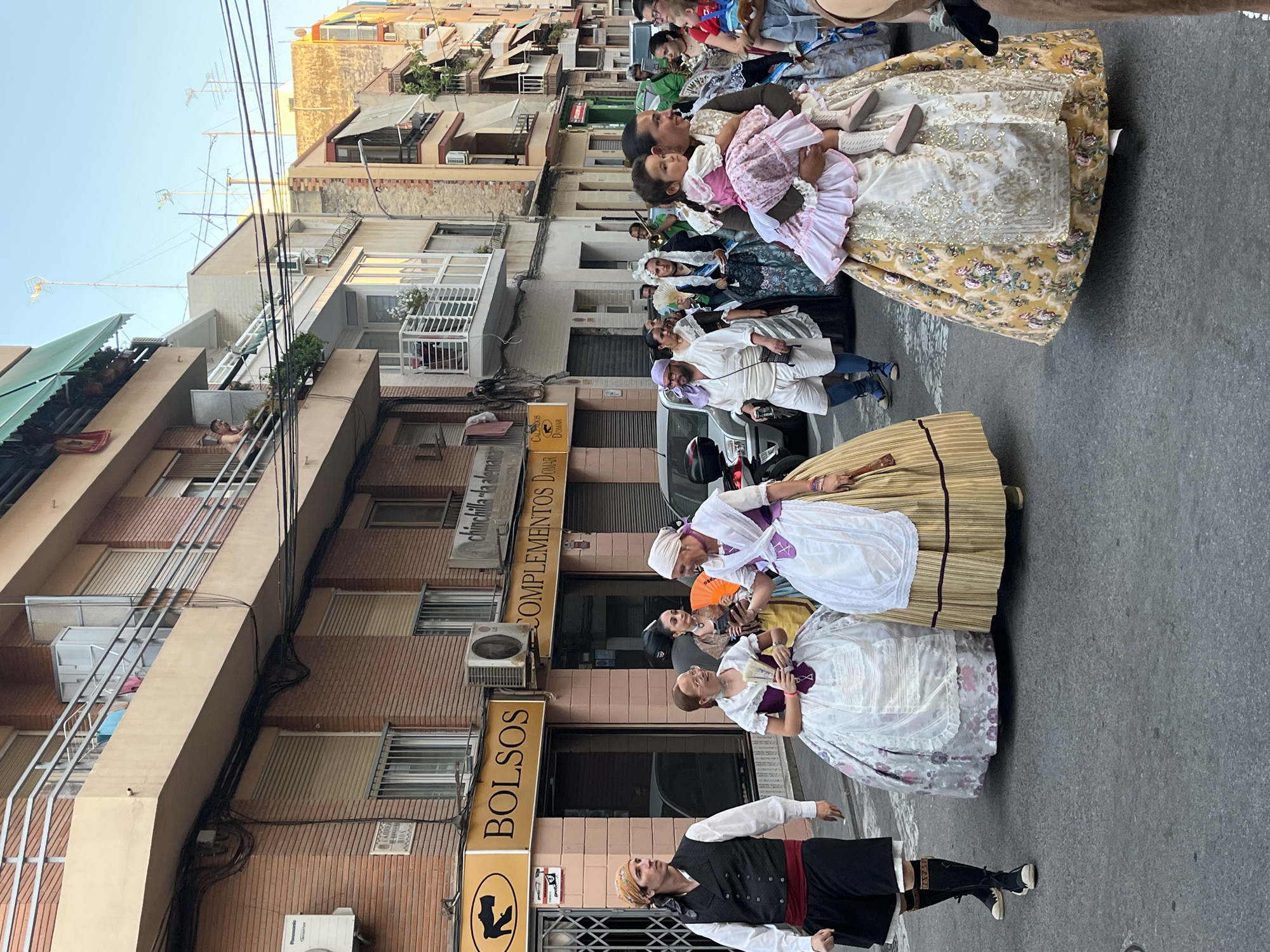 This media file captures a vibrant street scene in Alicante, Spain, likely during a festival or cultural event. A procession of people, many dressed in traditional Valencian attire, walk down a narrow street lined with multi-story buildings. The attire of the women includes long, elaborate skirts with floral patterns and embroidered bodices, often paired with white aprons and headscarves. Some individuals are holding fans and what appear to be bouquets of flowers. A man in the foreground wears a black vest over a white shirt, a red sash, dark pants, and a headscarf.
The architecture suggests a Mediterranean setting, with buildings featuring balconies, awnings, and what appear to be air conditioning units. The time of day is likely late afternoon or early evening, as indicated by the warm, golden light casting long shadows. The sky is a clear blue.
Visible text on storefronts includes "BOLSOS" (bags), "COMPLEMENTOS DOMAR" (Domar accessories), and "Chinchilla da la Alegría" (Chinchilla of Joy). The overall atmosphere appears festive and celebratory, with a sense of community and cultural pride.