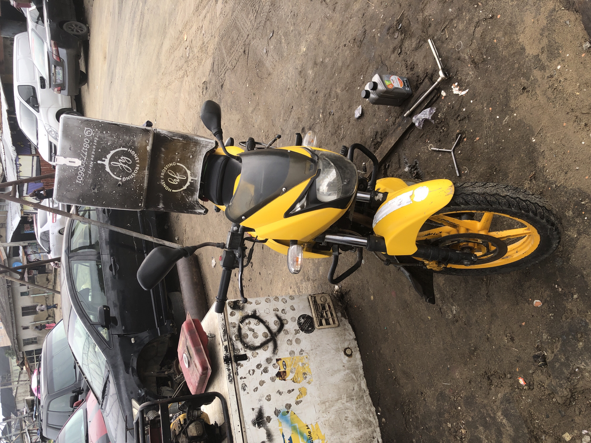 The image displays a bright yellow motorcycle with a prominent black delivery box mounted on its rear, parked on a dirt ground. The delivery box features white branding including a circular logo with stylized 'hf' initials and the words "HONEY FROSTING." A phone number, "08177306601," and the slogan "Eat and Do More" are also visible on the box.
The immediate surroundings indicate a repair or maintenance area. Various tools, including a silver socket wrench and a Y-shaped wrench, are scattered on the dirt near the motorcycle's front wheel, along with two plastic bottles of motor oil. The motorcycle itself has yellow rims, black handlebars, and a black windscreen.
In the background, several vehicles are parked. A black sedan with its front right wheel removed is visible to the left of the motorcycle, and a white panel van is parked further back. The ground is unpaved, covered in dirt with visible tire tracks and some debris. A red portable fuel tank rests on a metal platform beside the motorcycle. The scene takes place during the daytime, under bright lighting, suggesting an outdoor environment in Ado, Nigeria.
