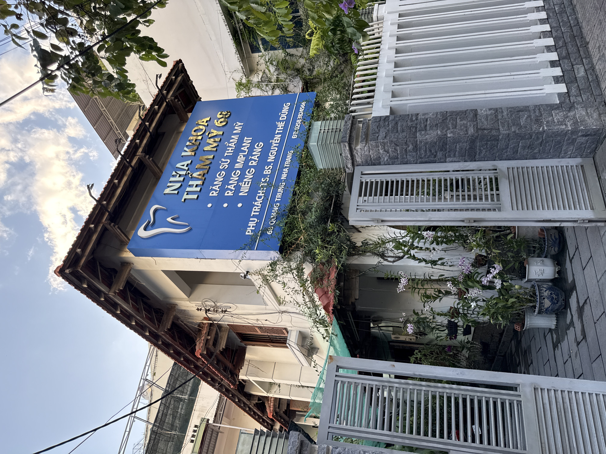 The image shows the exterior of a dental clinic named "NHA KHOA THẨM MỸ 69" located at 69 Quang Trung, Nha Trang, Vietnam. The clinic is situated in a building with a white facade and a traditional tiled roof. A prominent blue sign with white text and a stylized tooth logo displays the clinic's name and services, which include "RĂNG SỨ THẨM MỸ", "RĂNG IMPLANT", and "NIỀNG RĂNG". Below this, it states "PHỤ TRÁCH: TS.BS. NGUYỄN THẾ DŨNG", along with the address and a phone number "ĐT: 02583824566".
The clinic is surrounded by lush greenery, with plants and flowers, including orchids in pots, growing around the entrance. A white metal fence with a stone pillar encloses the property. The scene is set outdoors, with a clear sky and some electrical wires visible overhead. The overall impression is of a well-established dental practice in a pleasant, vegetated urban environment.