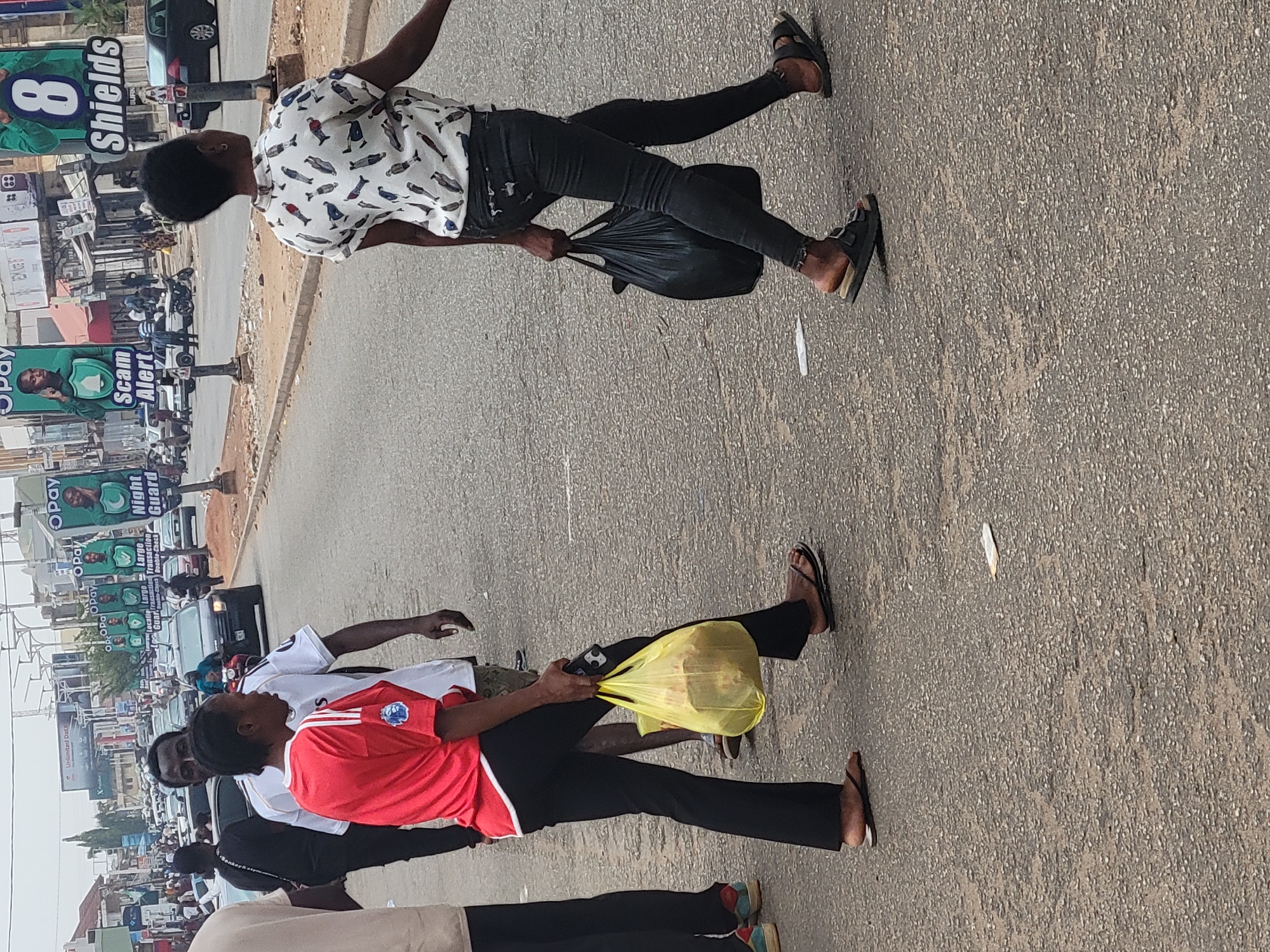 The image depicts a street scene in Jos, Nigeria, characterized by pedestrian activity and urban features. Two individuals are walking along the asphalt street, each carrying a bag: one a black plastic bag and the other a yellow plastic bag. The person with the black bag wears a white, patterned shirt and black pants, while the other wears a red and white sports jersey and black pants. They both wear open-toed shoes. In the background, a row of commercial billboards and signs lines the edge of the street. These signs advertise services such as 'Opay,' 'Night Guard,' and caution against scams, with the name 'Shields' appearing prominently on a large sign. Cars and motorcycles are visible along the street, indicating active traffic.
