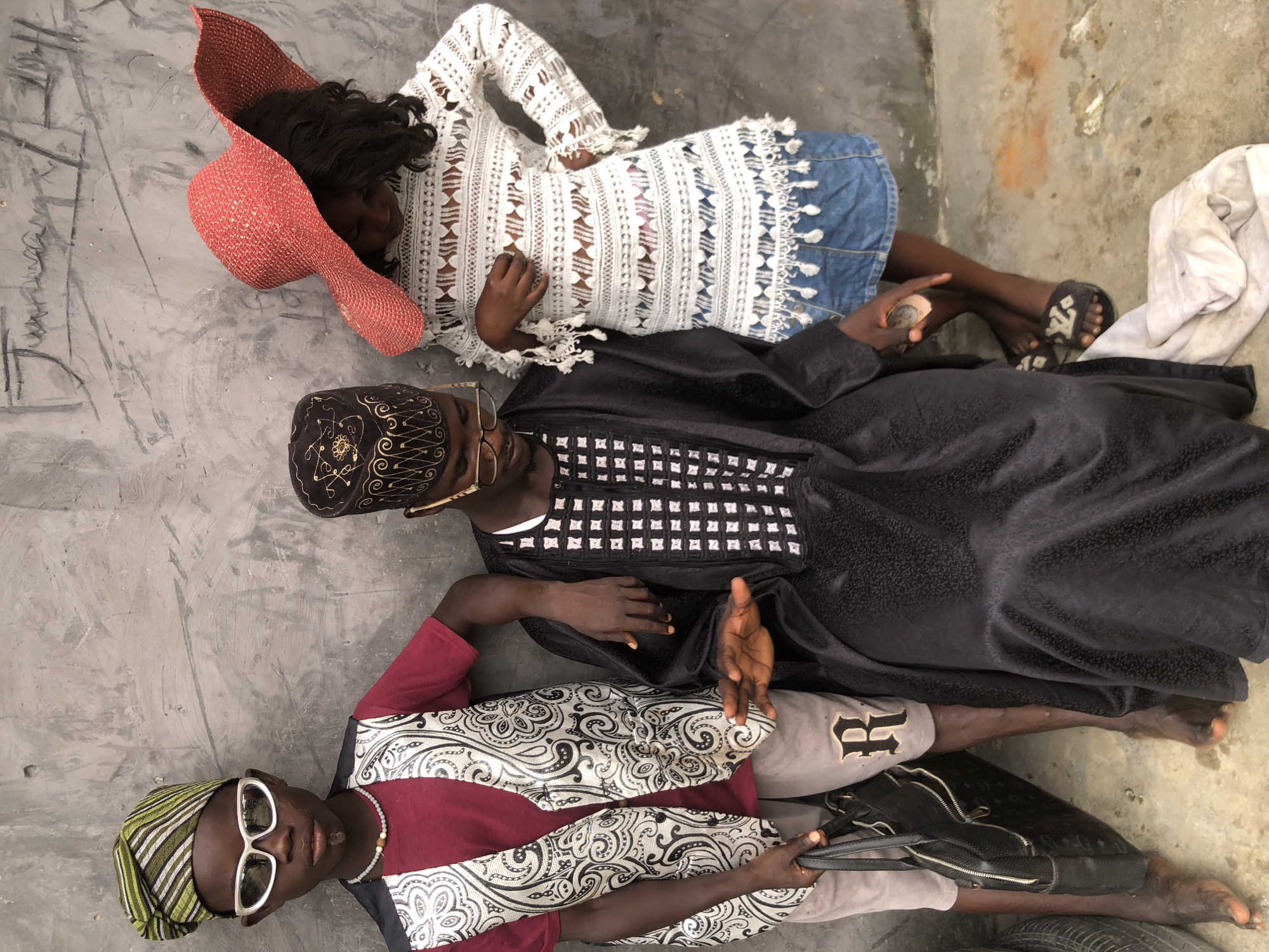 A medium shot captures three children posing against a textured grey wall in Ado, Nigeria. On the left, a boy wearing a striped green and white headwrap, white sunglasses, a maroon t-shirt, and a patterned vest stands with a black handbag. In the center, a boy dressed in a black kaftan with white square patterns and a patterned cap sits, leaning forward with his hands outstretched. On the right, a girl with a wide-brimmed red hat wears a white lace top and a denim skirt. The children are positioned close together, suggesting a shared moment or photoshoot. The environment appears to be an outdoor urban setting, indicated by the rough wall and the presence of what looks like a tire on the left edge of the frame. The overall mood is one of posed portraiture, with each child displaying a distinct style and demeanor.