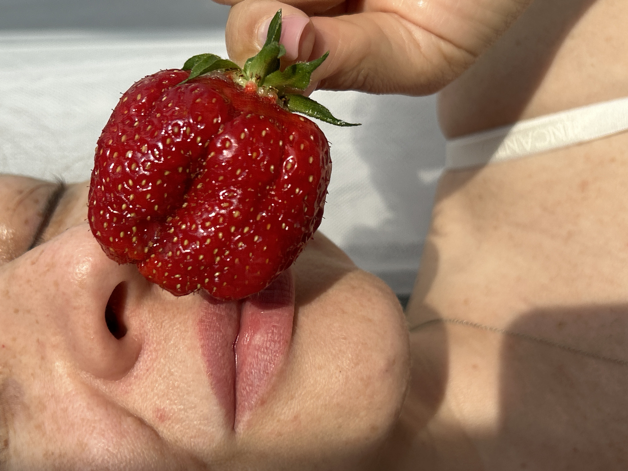A close-up, medium shot focuses on a woman's face and a large strawberry held near her mouth. The woman's nose and lips are visible, with a smattering of freckles across her skin. Her lips are slightly parted, and her mouth is positioned as if she is about to eat or taste the strawberry. The strawberry is bright red with visible seeds and a green stem. Her hand, with neatly manicured fingernails, holds the strawberry. In the background, a white, slightly blurred object, possibly a wall or curtain, is visible, with harsh sunlight casting shadows. The woman's shoulder and the strap of a white garment are also visible, hinting at an indoor or outdoor setting with strong lighting. No specific location in Chaikovsky, Russia can be determined from the visual information provided in the image.