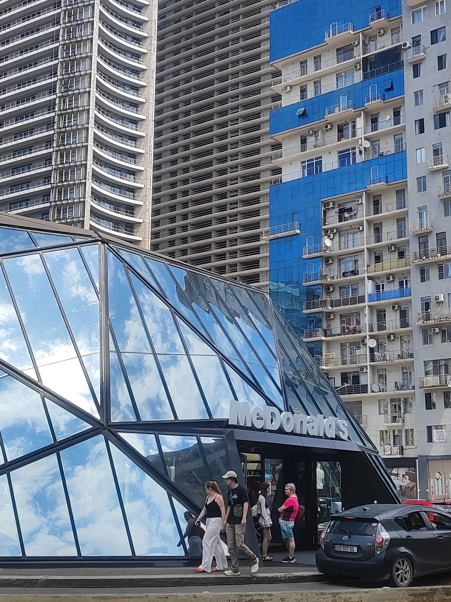 The image captures a street-level view of a modern McDonald's restaurant in Batumi, Georgia. The restaurant features a striking, angular glass facade that reflects the sky and surrounding buildings. Behind the McDonald's, several tall residential buildings are visible. One building immediately behind the McDonald's is characterized by its bright blue glass exterior, contrasting with the grey facade of an adjacent, older building. Further in the background, a skeletal building, likely under construction, displays rows of unfinished floors and balconies.
In the foreground, a dark-colored hatchback car is parked on the right side of the street, facing away from the viewer. A few people are walking on the sidewalk in front of the McDonald's. One person, wearing a black tank top and white pants, is walking to the left, with another person, possibly a man in a t-shirt and cap, walking alongside. Two other individuals, one in a pink top and patterned shorts, and another in a light-colored outfit, are standing near the entrance of the McDonald's. The scene appears to be set during the daytime, with natural light illuminating the area. The presence of multiple tall buildings suggests an urban environment, consistent with a city like Batumi.