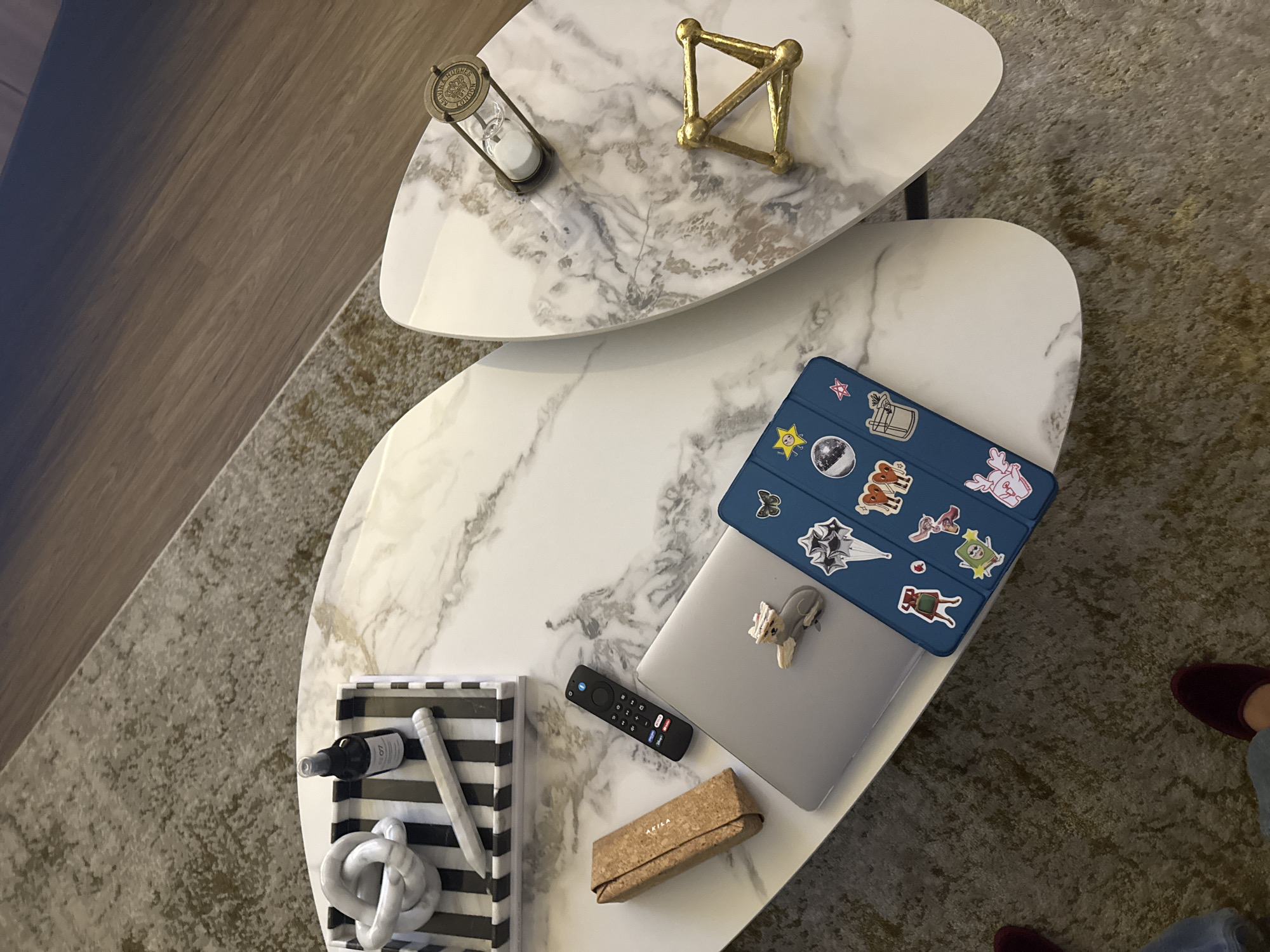 The image shows an overhead view of a two-tiered coffee table with a faux marble finish, likely located in a living room setting. The top tier features an hourglass and a geometric gold-colored object. The lower tier holds a laptop with a blue case adorned with stickers, a television remote, a cork box, and a decorative tray containing a bottle, a pencil, and a white knot sculpture. The surrounding environment includes a beige patterned rug and a glimpse of flooring. There is no evidence to determine the location within the city of San Bernardino Tlaxcalancingo, Mexico.