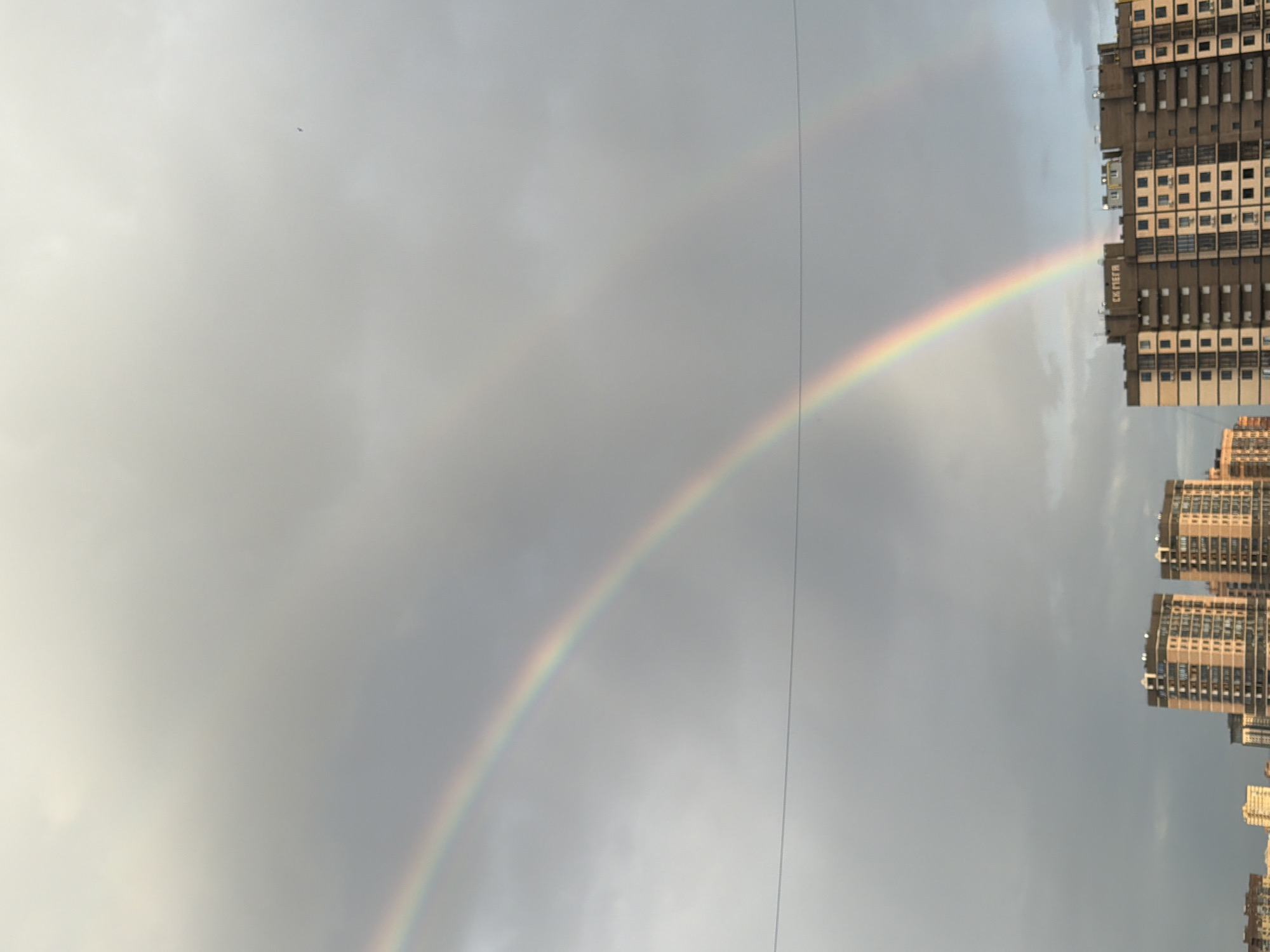 The image shows a double rainbow over a cloudy sky, with a row of apartment buildings on the right side. The buildings appear to be located in an urban area, likely in Ovoshchnoy, Russia, based on the signage visible on the buildings. A power line cuts through the middle of the rainbow. A small bird is flying in the sky. The rainbow's arc extends across the sky from left to right.