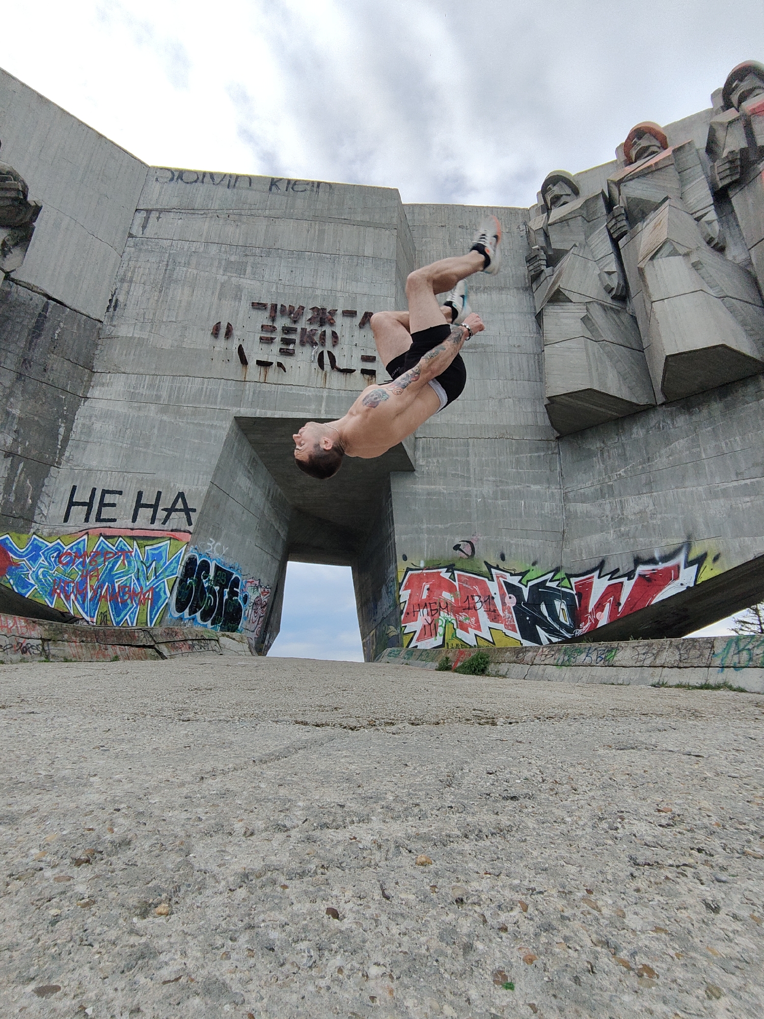 A man is performing a backflip in front of the Buzludzha Monument in Bulgaria. The man is shirtless and wearing black shorts and sneakers. The monument is large, concrete, and decorated with graffiti. The sky is cloudy.