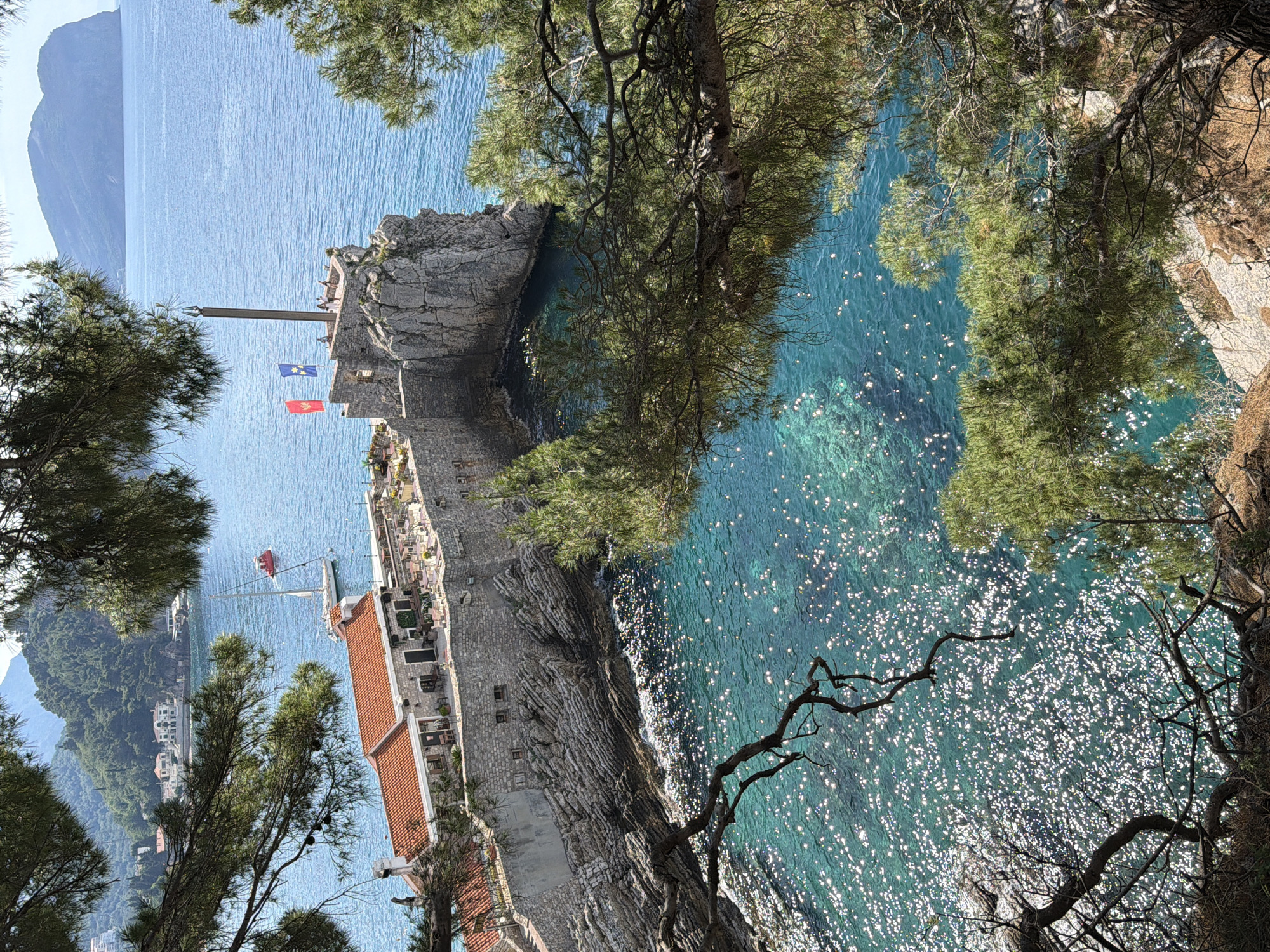 The image, taken from an elevated position, presents a coastal view in Petrovac centar, Montenegro. The sea is a prominent feature, shimmering with reflected sunlight. Part of a stone structure, possibly a fortress or historical building, juts out into the water, displaying walls and an orange tiled roof. A flagpole stands atop the structure, displaying two flags. Lush green trees frame the view, with branches obscuring parts of the scene. In the background, a mountainous coastline is visible. A sailing vessel is visible in the water next to the tiled roof structure.