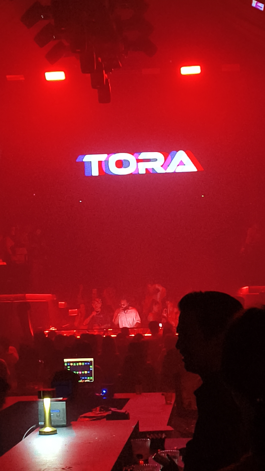 In North Kuta, Indonesia, a dimly lit nightclub scene unfolds, dominated by a vibrant red glow from overhead stage lighting. The word "TORA" is emblazoned in large, colorful lettering across a screen at the back of the venue, with blue and white hues creating a striking contrast against the red. Below the screen, two individuals, likely DJs, stand behind a console, their figures silhouetted and illuminated by the ambient light. A crowd of people can be seen gathered in front of the stage, their faces obscured by the low lighting and distance. In the foreground, a bar counter is visible, adorned with various objects including a monitor displaying colorful graphics, a golden-hued lamp, and several glasses. The overall atmosphere suggests a lively night at a music venue, with the red lighting creating an intense and energetic mood.