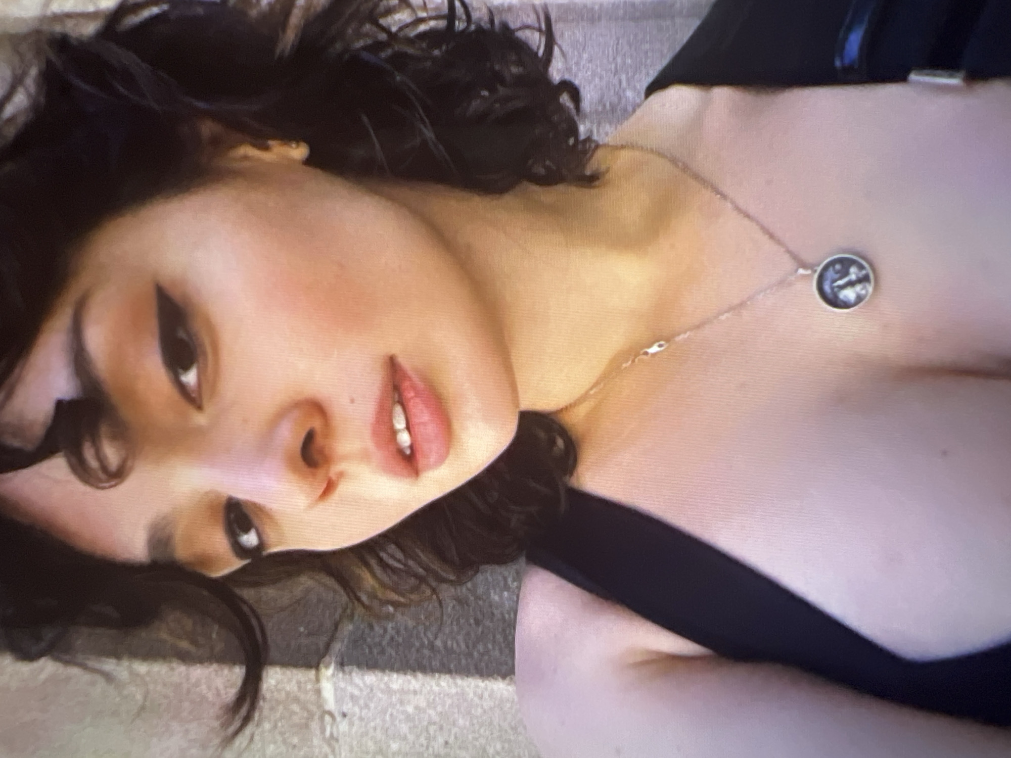 A close-up, chest-up selfie of a young woman with dark, curly hair and light skin is shown. She is wearing a black top and a silver necklace with a circular pendant. Her makeup includes eyeliner and blush. The background appears to be an outdoor setting with a textured, light-colored wall and some shadows. There is no visible connection to Pyatigorsk, Russia in the image.