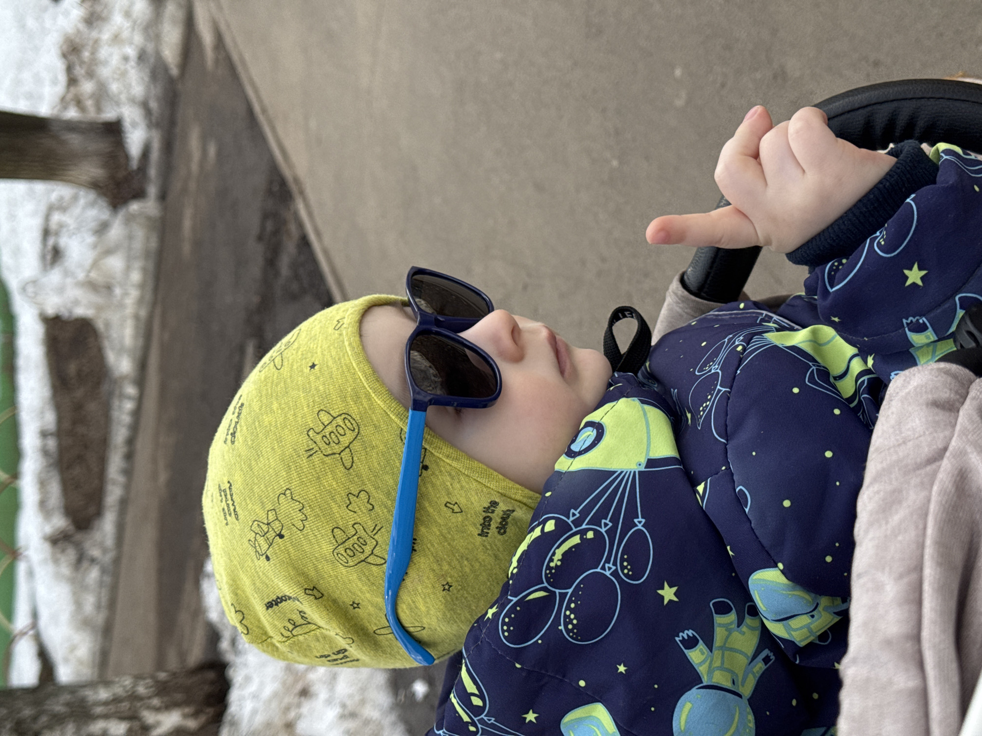 A young child wearing a yellow hat with airplane prints and blue sunglasses is sitting in a stroller outdoors. The child is wearing a navy blue jacket with a space-themed pattern. The stroller handle is black and the child's hand is resting on it. The background shows an urban environment, with snow on the ground and a concrete structure behind the child. There is no specific evidence in the image to pinpoint the location in Izhevsk, Russia.