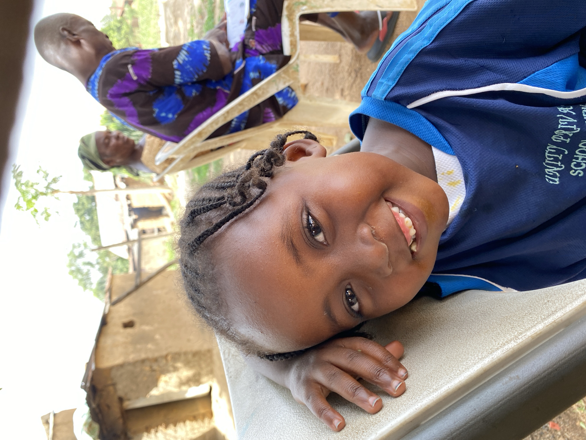 A young African girl with braided hair, wearing a blue school uniform with the words "UNITY PRIVA SCHOOL" visible, is lying on a table with her head and hand resting on the surface. She is smiling. In the background, two adults are seated on chairs. One adult is wearing a brown and blue patterned garment. The background shows a sunlit outdoor scene with a partially visible building, suggesting a rural or less developed area in Kuru Dungus, Nigeria.