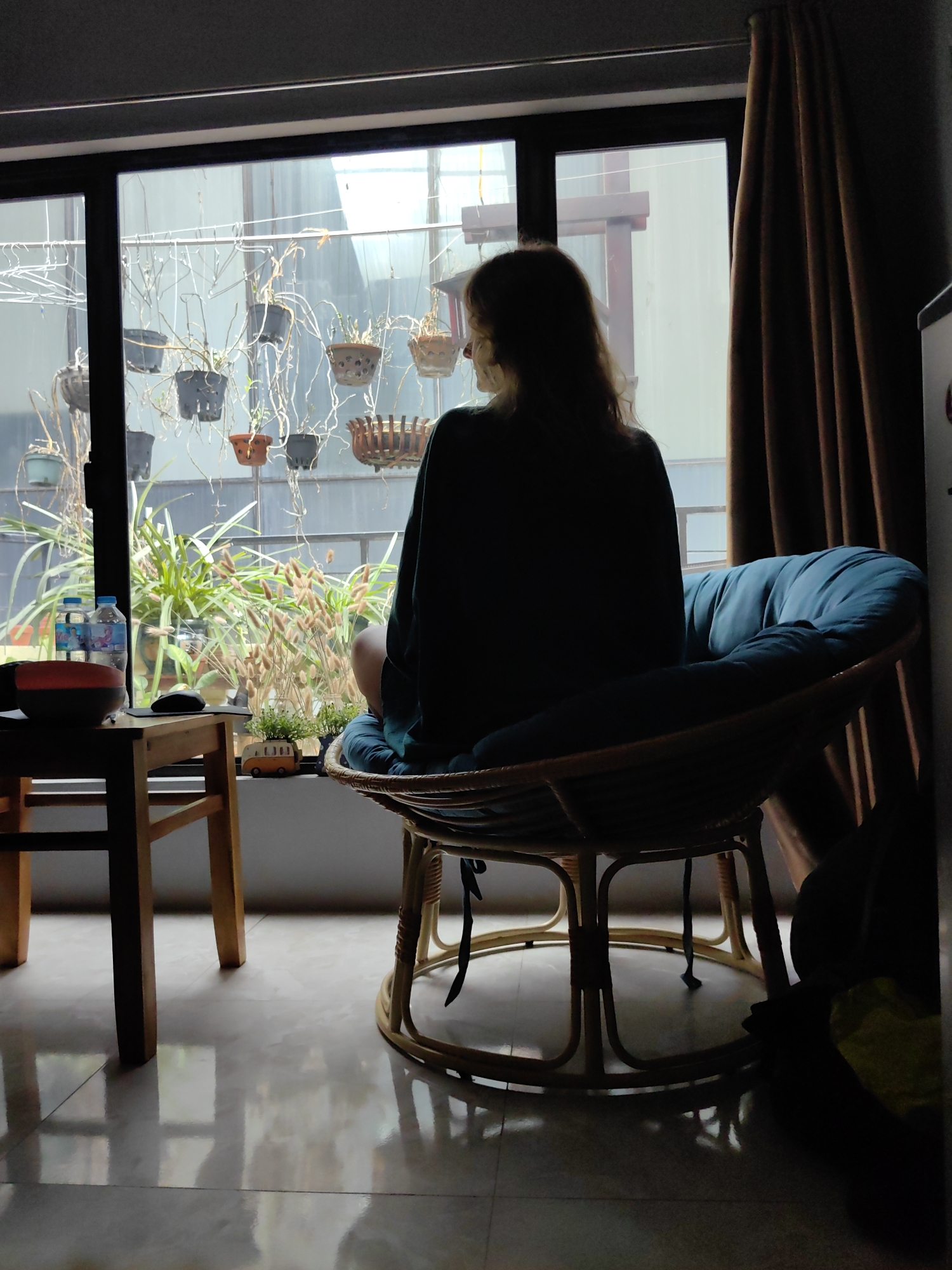 The image depicts the interior of a room, presumably in Hà Nội, Vietnam, as the style of the architecture and hanging potted plants visible through the window are characteristic of the region. A person, possibly female with long hair, is seated in a papasan chair positioned in front of a large window. The person is facing away from the camera, obscuring most of their features. The window looks out onto a space where various plants, some in hanging pots, are suspended from a structure. The external view beyond the plants shows a building under construction, which is typical of the rapid urban development in Vietnamese cities. A small wooden table stands to the left of the chair, holding items including water bottles. The room appears to be well-lit by natural light coming through the window. Brown curtains hang to the right of the window, partially blocking a view. The floor is tiled and reflective, indicating a hard surface.