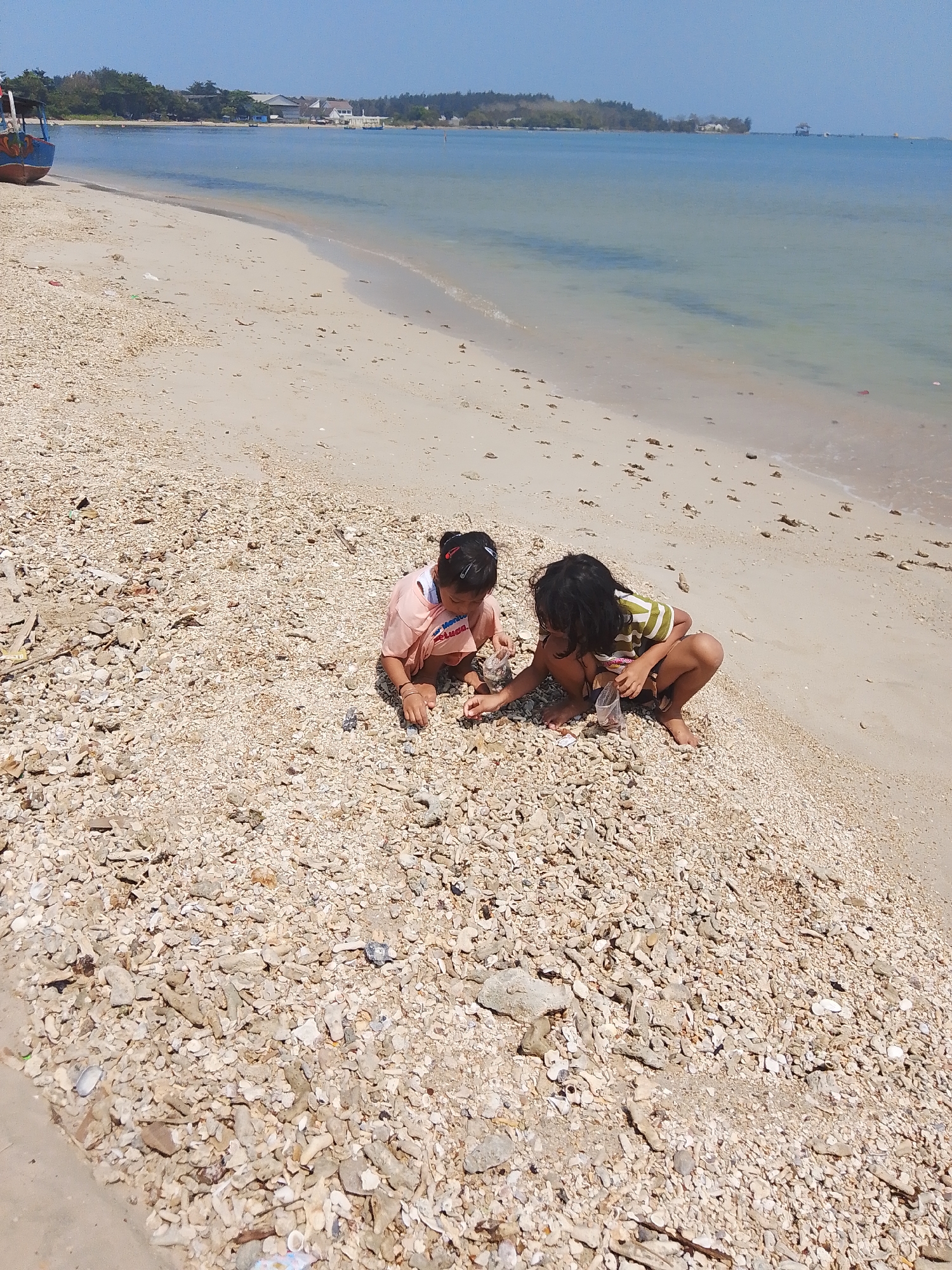 Two young children are crouched on a beach covered in shells and rocks in Tegalsambi, Indonesia. One child, on the left, wears a peach-colored shirt and has dark hair pulled back in a ponytail. The other child, on the right, wears a green and white striped shirt and has dark, long hair. Both children are looking down at the shells and appear to be collecting or examining them. The beach is sandy with a high concentration of shell fragments and small rocks, sloping down to the shallow, calm ocean. In the background, there is a line of trees and some buildings on the coastline under a clear blue sky. A partially visible blue and red boat is docked on the left side of the image, near the edge of the water.