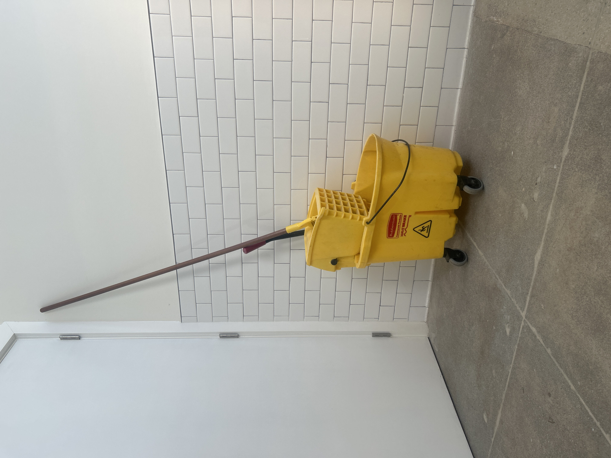 The image depicts a yellow mop bucket with a mop leaning against a wall with white rectangular tiles above a white door. The scene appears to be inside a building, likely a commercial or institutional space, in Los Angeles, United States of America. The floor is a light grey, tiled surface. There are no people visible in the image.