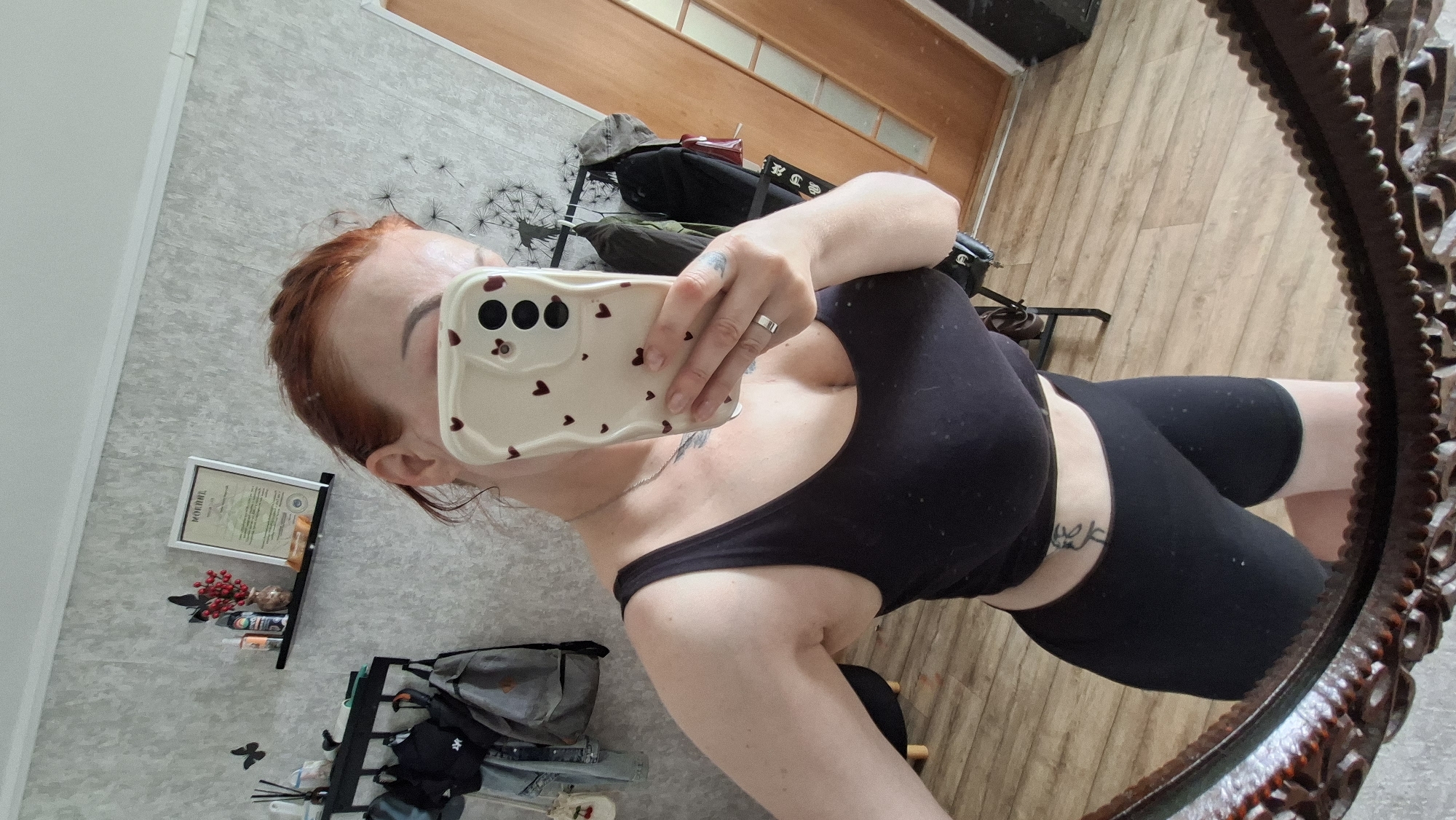 A woman with red hair is taking a mirror selfie indoors. She is wearing a black tank top and black cycling shorts. She has tattoos on her arm and torso. The background shows a wall with a shelf holding a certificate and some decorative items, as well as a rack of clothing. The floor is made of wood. The image is not consistent with the city of Myadzyel, Belarus, as it appears to be taken in a residential interior.