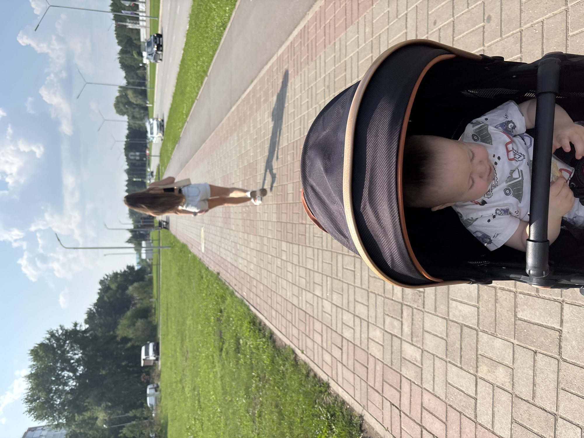 A baby is in a stroller on a paved sidewalk. The stroller has a black canopy with an orange trim. The baby is wearing a white t-shirt with a pattern of cartoon vehicles. In the background, a young woman with long brown hair is walking away from the stroller on the same sidewalk. She is wearing a black crop top and light-colored denim shorts. Beyond the sidewalk is a grassy area and a road with cars driving on it. Streetlights are visible along the road. The scene appears to be in an urban environment, likely Naberezhnye Chelny, Russia, given the context. The sky is blue with scattered white clouds, indicating a sunny day.
