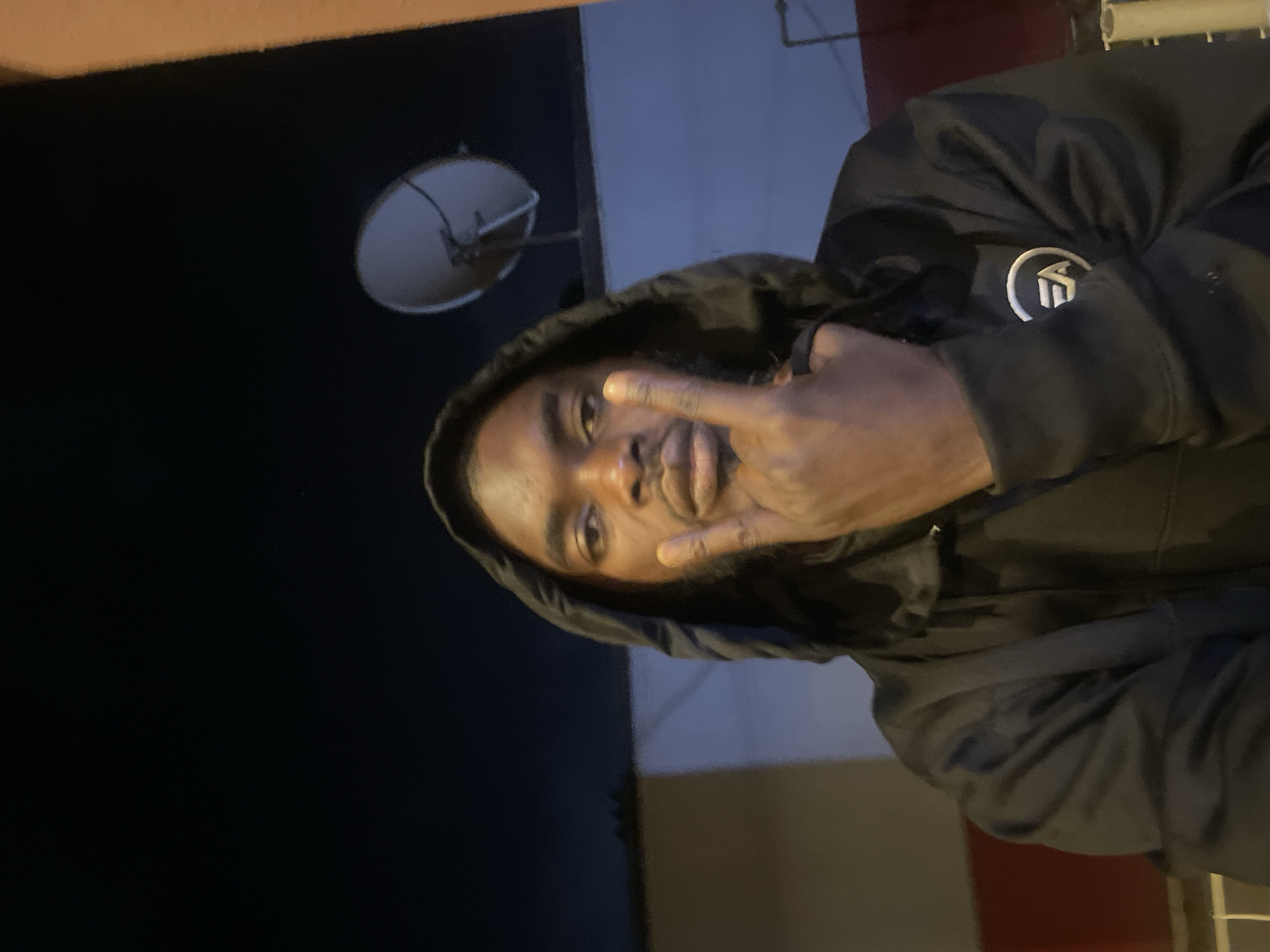 A dark-skinned man wearing a black hooded sweatshirt is making a hand gesture with his right hand, holding up his index and pinky fingers. He is looking at the camera. Above his head and to the left is a satellite dish affixed to a building. It appears to be night or dusk. The exact location within the city of Jos, Nigeria cannot be determined from the image.