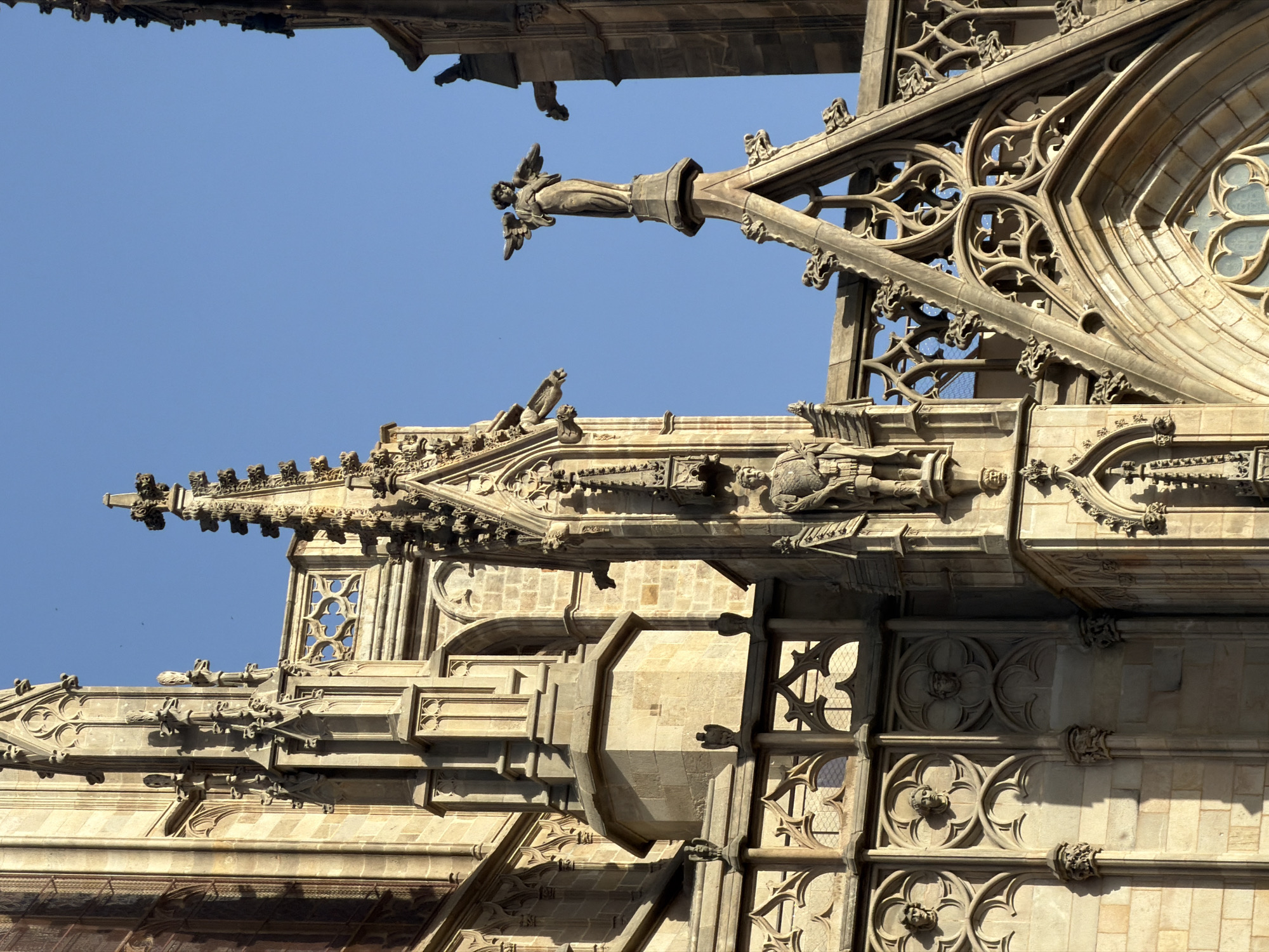 The image shows a close-up view of the exterior of the Barcelona Cathedral, located in the Gothic Quarter of Barcelona, Spain. The architecture is characterized by Gothic style, with intricate stone carvings, pointed arches, and spires. Several statues are visible, including one of an angel on a tall pinnacle and another of a figure in armor on a projecting section of the building. Gargoyles are also present, projecting from the stonework. The facade is constructed from light-colored stone, and the clear blue sky serves as the backdrop. Sunlight casts shadows on the architectural details, highlighting the textures and depths of the carvings.