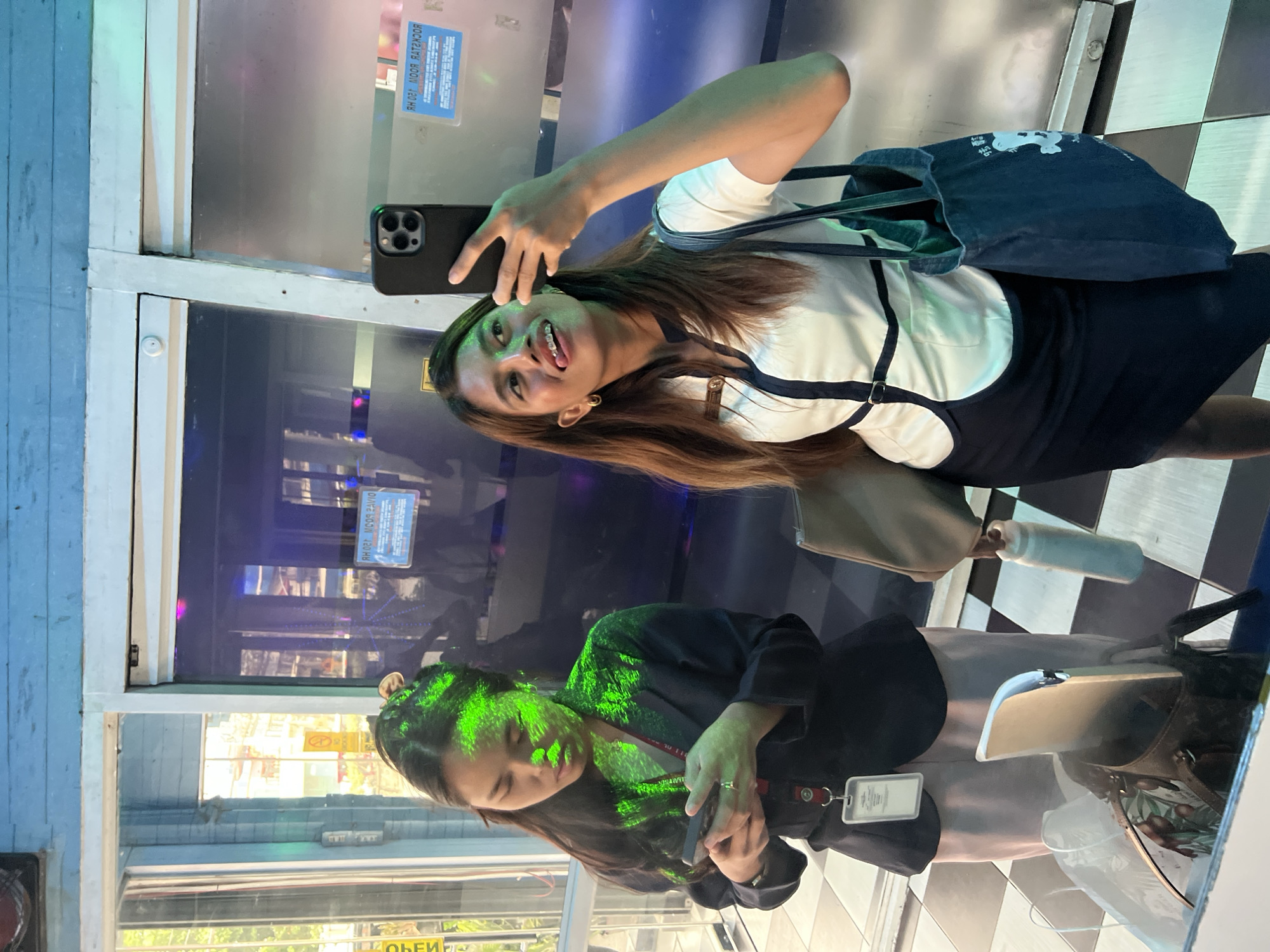 The image shows a vertical mirror reflection of two young women. The woman on the left is facing the viewer and taking a selfie with a smartphone. She is wearing a white shirt with a dark pinafore dress, a denim tote bag slung over her shoulder, and has braces. The woman on the right is looking down at her phone, with bright green light patterns projected onto her face and hair. She is wearing a dark jacket over a light-colored top, and a lanyard with an ID card.
The setting appears to be an indoor space with a checkered floor and reflective surfaces, possibly a shopping mall or an entertainment venue. The background shows distorted reflections of what seems to be a brightly lit interior with colorful lights. The time of day is not discernible from the image, nor is the weather.
The woman on the left's expression is playful and she is sticking out her tongue, suggesting a lighthearted or fun mood. The woman on the right's expression is more subdued, focused on her phone.
Visible text includes "ROCKSTAR BOOM 120 HR" on a blue sticker reflected in the mirror on the top left. Another reflection on the bottom left shows a yellow sign with text that is partially obscured but appears to say "OPEN". The ID card on the woman on the right has text on it, but it is too blurry to read. The denim bag has a white graphic on it.
The overall impression is a candid moment captured through a mirror, showcasing casual interactions and perhaps a shared outing.
