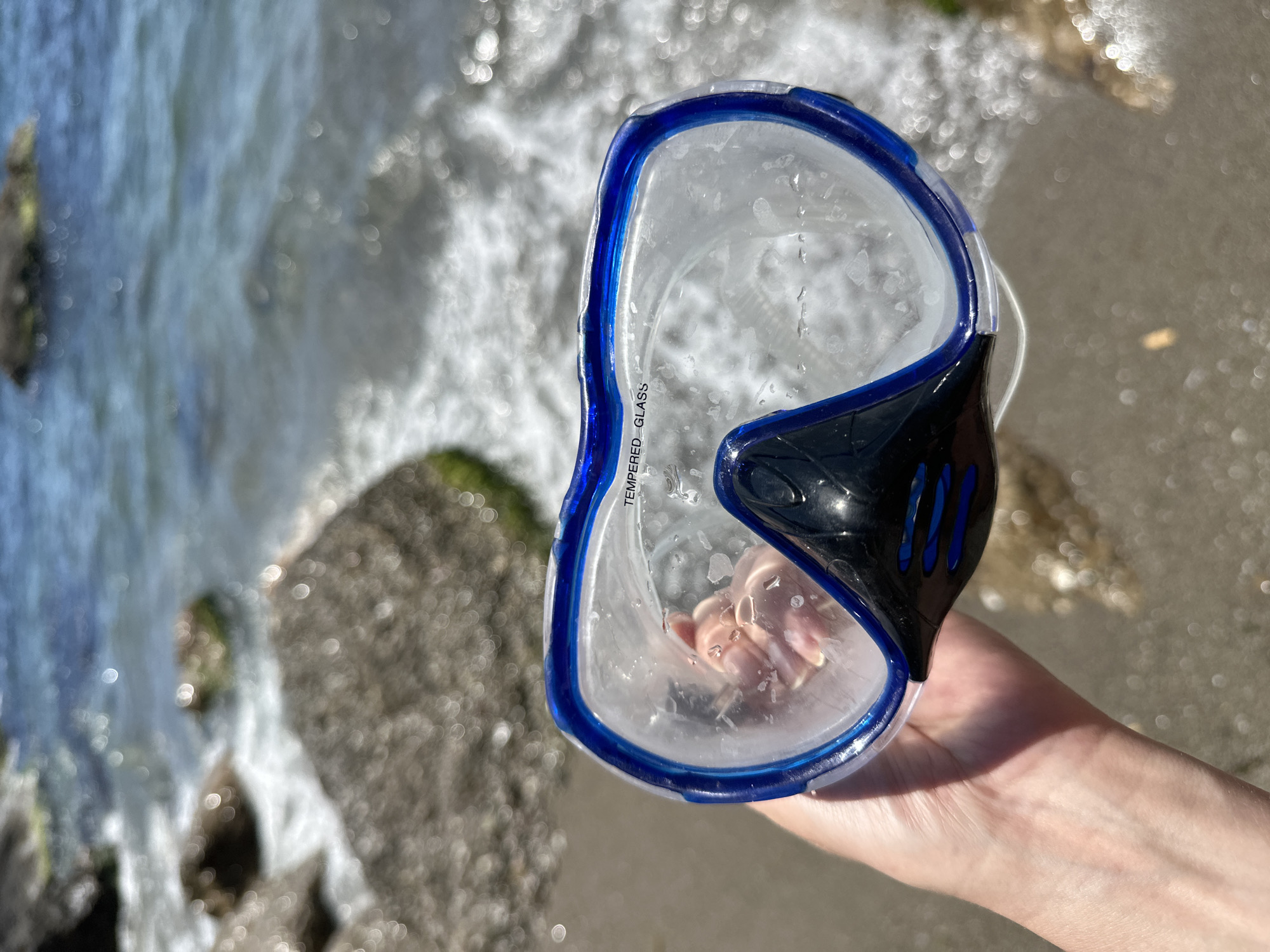 A person's hand holds a blue and black swimming mask in the foreground. The mask is wet and has water droplets on the inside of the lens. The background shows a rocky shoreline with waves crashing on the beach. The water is a clear blue, and the sky is bright. The location is the beach.