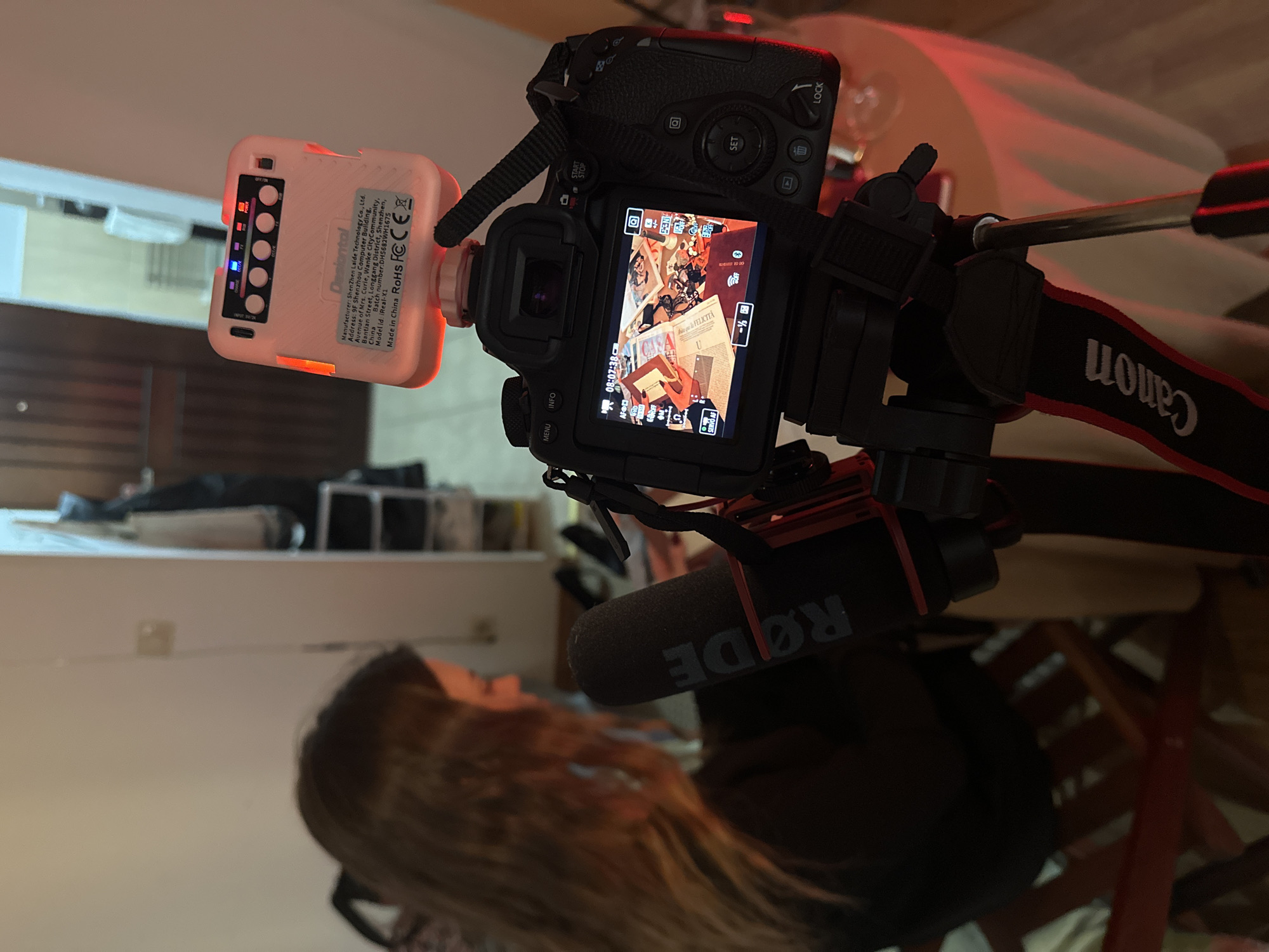 An indoor scene is captured within a residence in Šušanj, Montenegro. A black Canon camera, equipped with a "Canon" branded strap, is prominently positioned in the foreground, mounted on a tripod. Attached to the camera's hot shoe is a white Deonlaitel LED light panel, emitting red light, with its "POWER" and "RGB" indicators illuminated. A black RØDE microphone is affixed to the camera setup, facing towards the rear of the frame. The camera's rear screen displays a live view or recorded footage of an overhead arrangement of papers, photographs, and a wooden 'U' on a red surface, with a visible timestamp of "08:07:38."
In the midground, a person with long brown hair, wearing a dark garment, is seated on a wooden chair at a table covered with a white tablecloth. Their profile and upper back are visible as they face away from the camera's front. The table and immediate surroundings are bathed in a red ambient light, and a clear glass is situated on the table to the camera's right. The background features a doorway leading to an adjacent, brightly lit room with white walls. To the right of the doorway, a storage unit or coat rack is partially discernible.