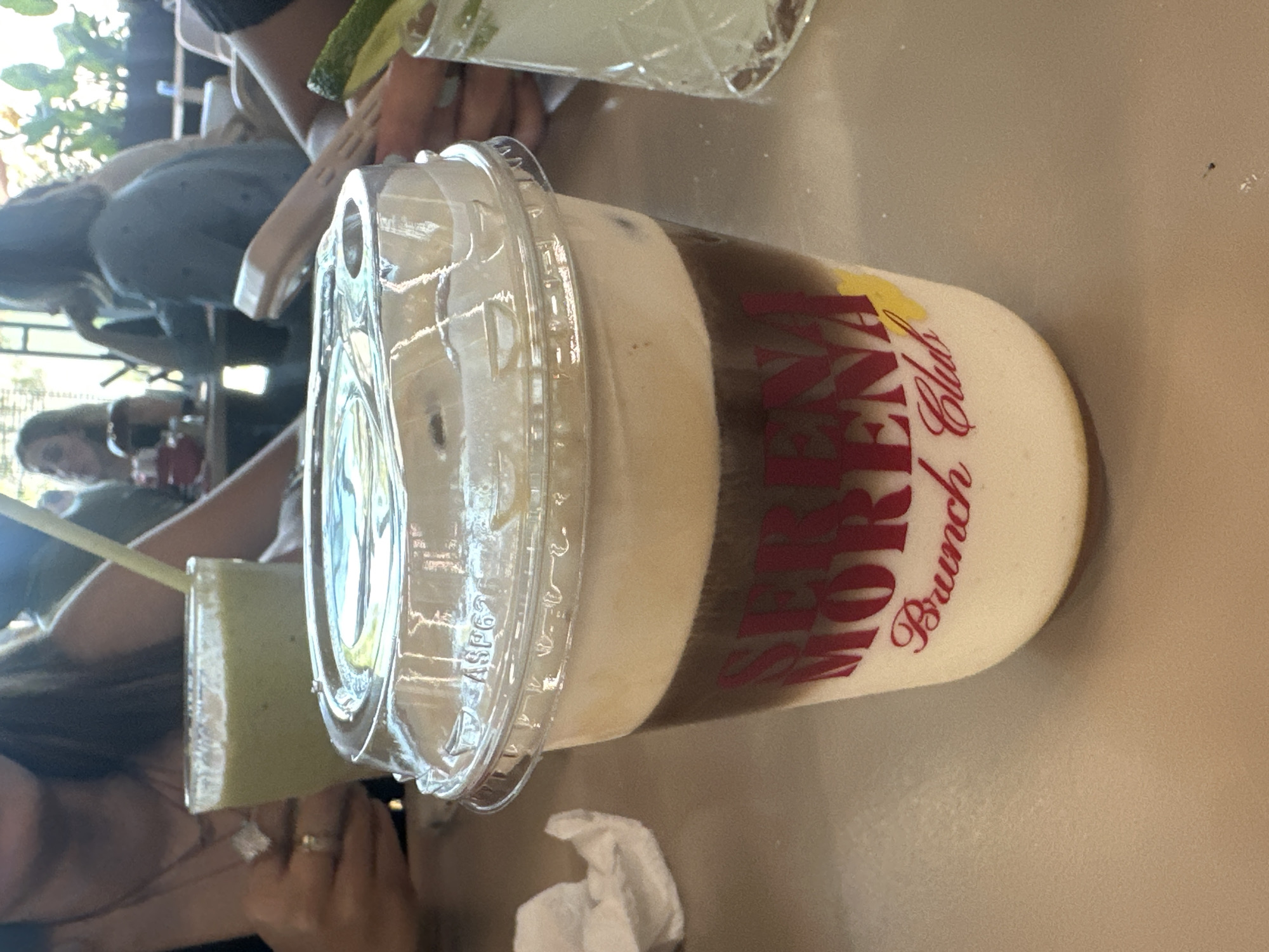 The image shows a group of people gathered at a table. The focus is on a tall, clear plastic cup filled with a layered beverage. The cup has a transparent plastic lid. The drink consists of a dark liquid, likely coffee, and a creamy white topping. The name "Serena Morena Brunch Club" is printed in red, stylized font around the cup. Next to it, there's another drink in a glass, featuring a green liquid with a straw. The table's surface is a neutral tan color. In the background, other individuals are partially visible, along with the surrounding environment of what seems to be an outdoor dining area, with plants and a glimpse of a building through the open side. There is an assumption that this location is in the city of Puebla, Mexico.