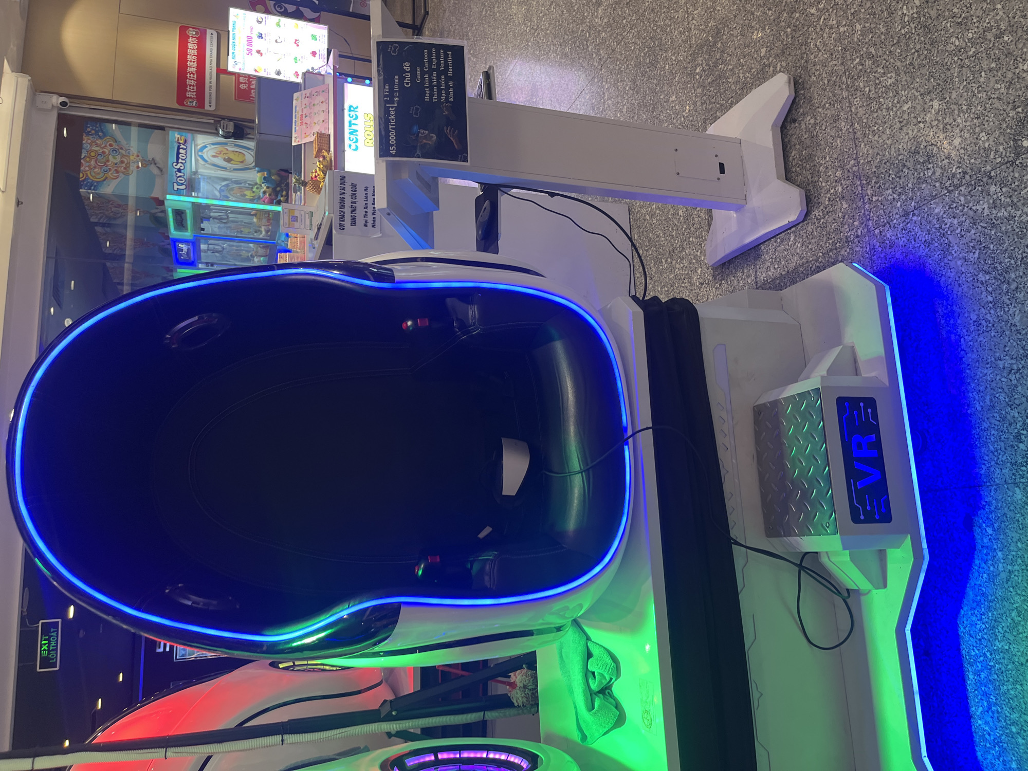 The image shows an indoor VR gaming station at Nha Trang Center, a shopping mall in Nha Trang Ward, Vietnam. The station features a large, egg-shaped seat with blue and green LED lighting. Signage indicates the games cost 45,000 VND per ticket, with options including cartoon, explore, venture, and horror themes. There are additional signs for other establishments such as "Center Rolls" and "Toy Story", along with a nail salon. The flooring has a gray speckled design.