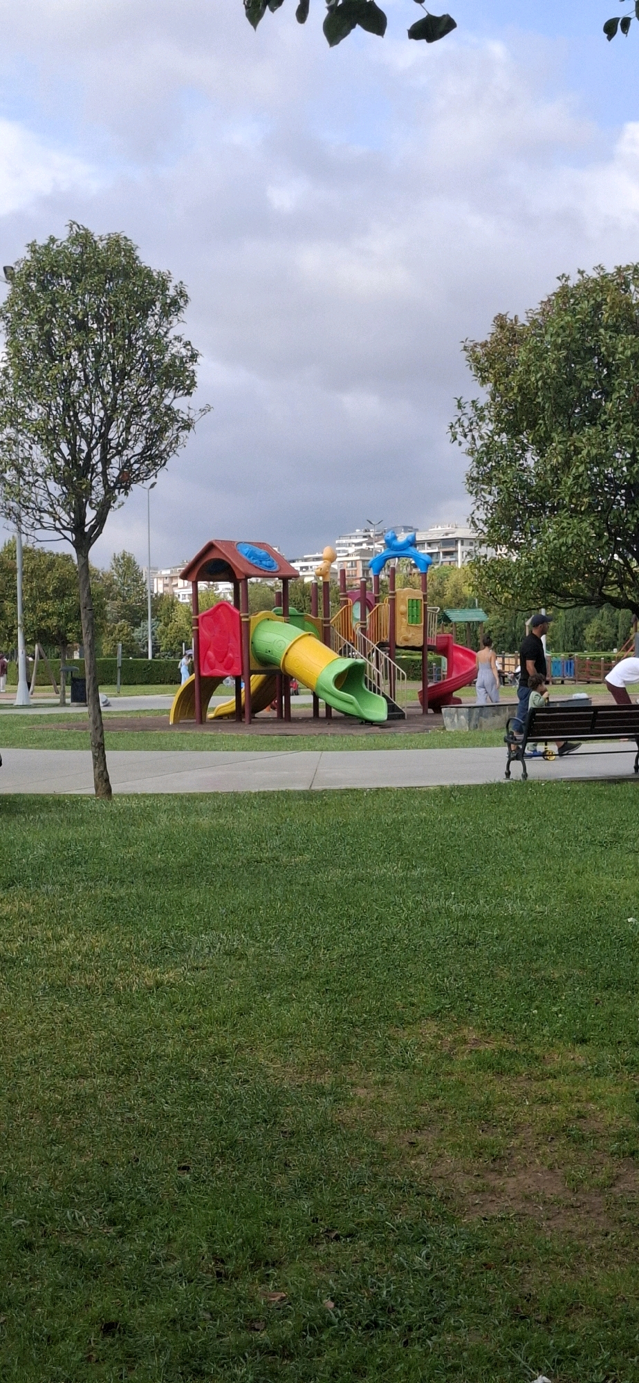 In Maltepe, Turkey, a public park features a vibrant children's playground with multiple slides and climbing structures. A red, yellow, and green tunnel slide is prominent, with a smaller red slide visible to its right. Behind the playground, trees and buildings form the background under a cloudy sky. In the foreground, a grassy lawn is dotted with a few people. A man in a black shirt and hat stands near a bench, looking towards the playground, while a child in a blue shirt stands beside him. Another person in a white shirt is partially visible on the right side of the frame.
