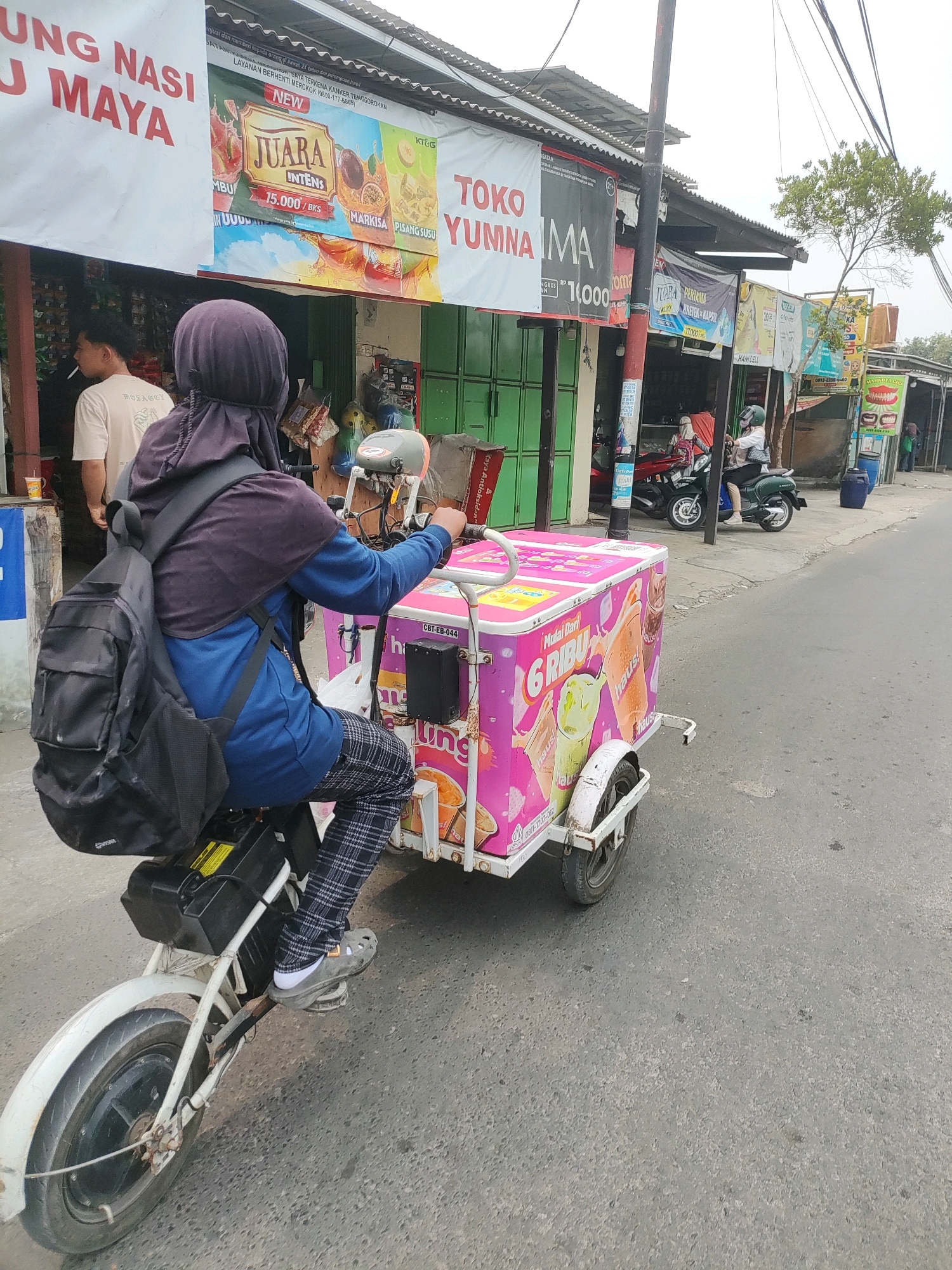 An adult, wearing a purple headscarf, blue jacket, and black and white checkered pants, rides an electric tricycle towing a pink ice cream cart. The tricycle has a black battery pack mounted on its frame. The ice cream cart is decorated with images of ice cream and text in Indonesian, including "Mulai Dari 6 RIBU" (Starting from 6 thousand). The person is seen from behind, and their right foot is on the pedal. The tricycle is moving down a paved road lined with small shops and banners. In the background, other shops are visible, as well as a person on a scooter and a blue trash bin. The scene appears to be in a commercial or residential area of Wanajaya, Indonesia, given the signage. The weather is overcast.
