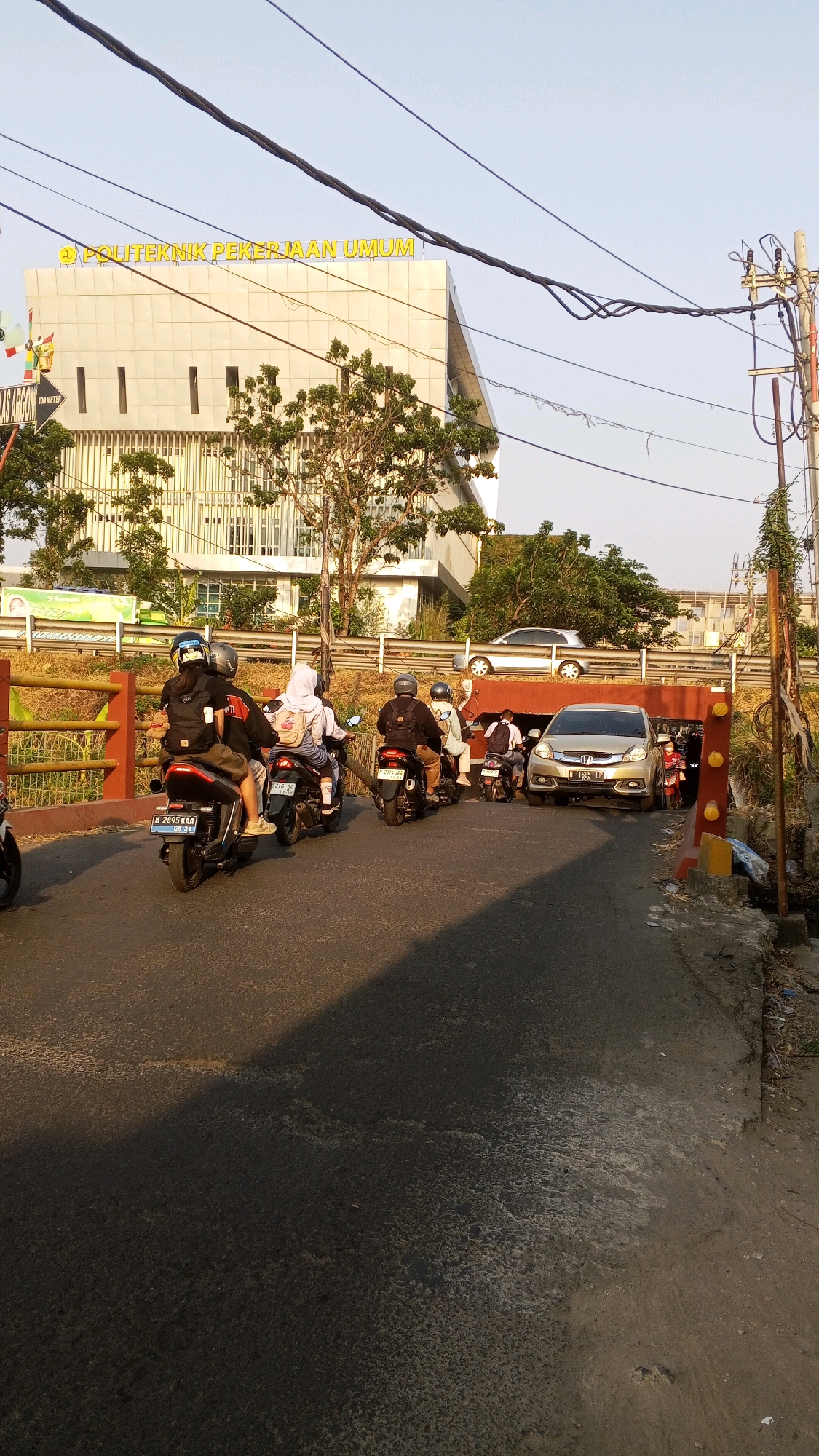 The image depicts a street scene in Semarang, Indonesia, near the "POLITEKNIK PEKERJAAN UMUM" (Public Works Polytechnic). A gold sedan is stopped in a tunnel-like underpass, its front license plate reading "H 1685 LP". Several motorbikes are lined up behind the car, some with passengers. On the left, three motorbikes are visible in the foreground, with riders wearing helmets and backpacks. The road surface appears wet and reflective in some areas, casting a shadow from the right side of the frame. In the background, a modern, light-colored building with the institution's name displayed in yellow lettering is prominently visible. Electrical wires and poles are present in the upper right corner of the image. The overall scene suggests a busy street during daylight hours, possibly during a traffic slowdown or waiting period.