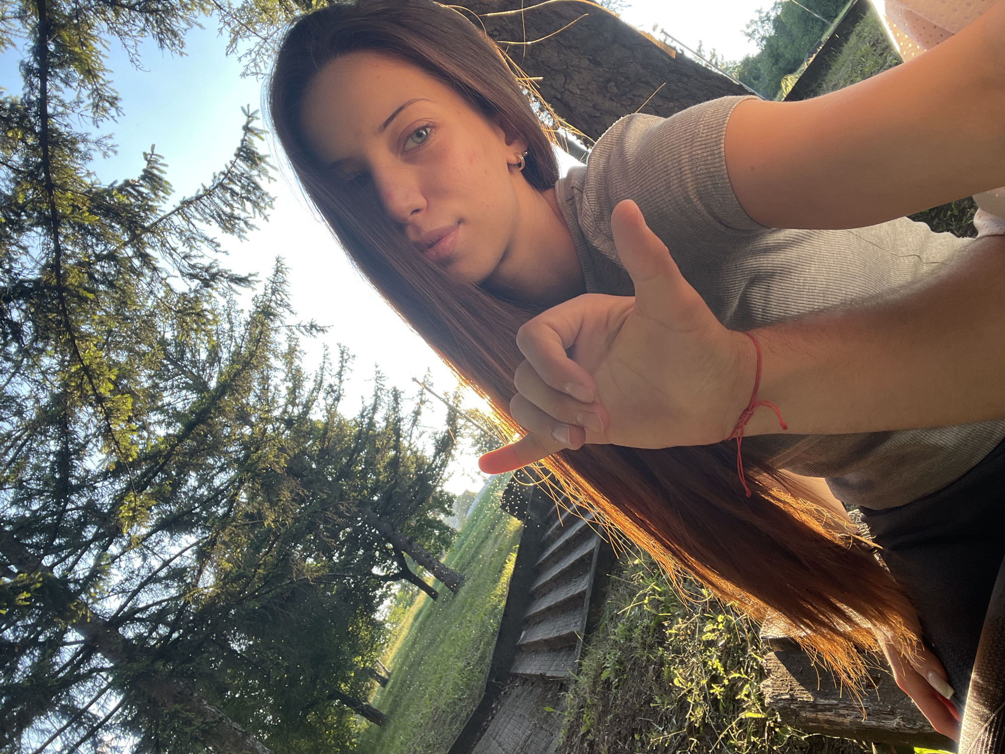 An adult female with long, brown hair is taking a selfie in an outdoor environment. She is wearing a grey, ribbed t-shirt and has a thin red bracelet on her left wrist. Her left hand is raised, forming a "shaka" sign with her thumb and pinky extended. Her eyes are open and looking towards the camera. She appears to be outdoors with trees and grass in the background, bathed in sunlight. The location is identified as Unknown, Unknown.